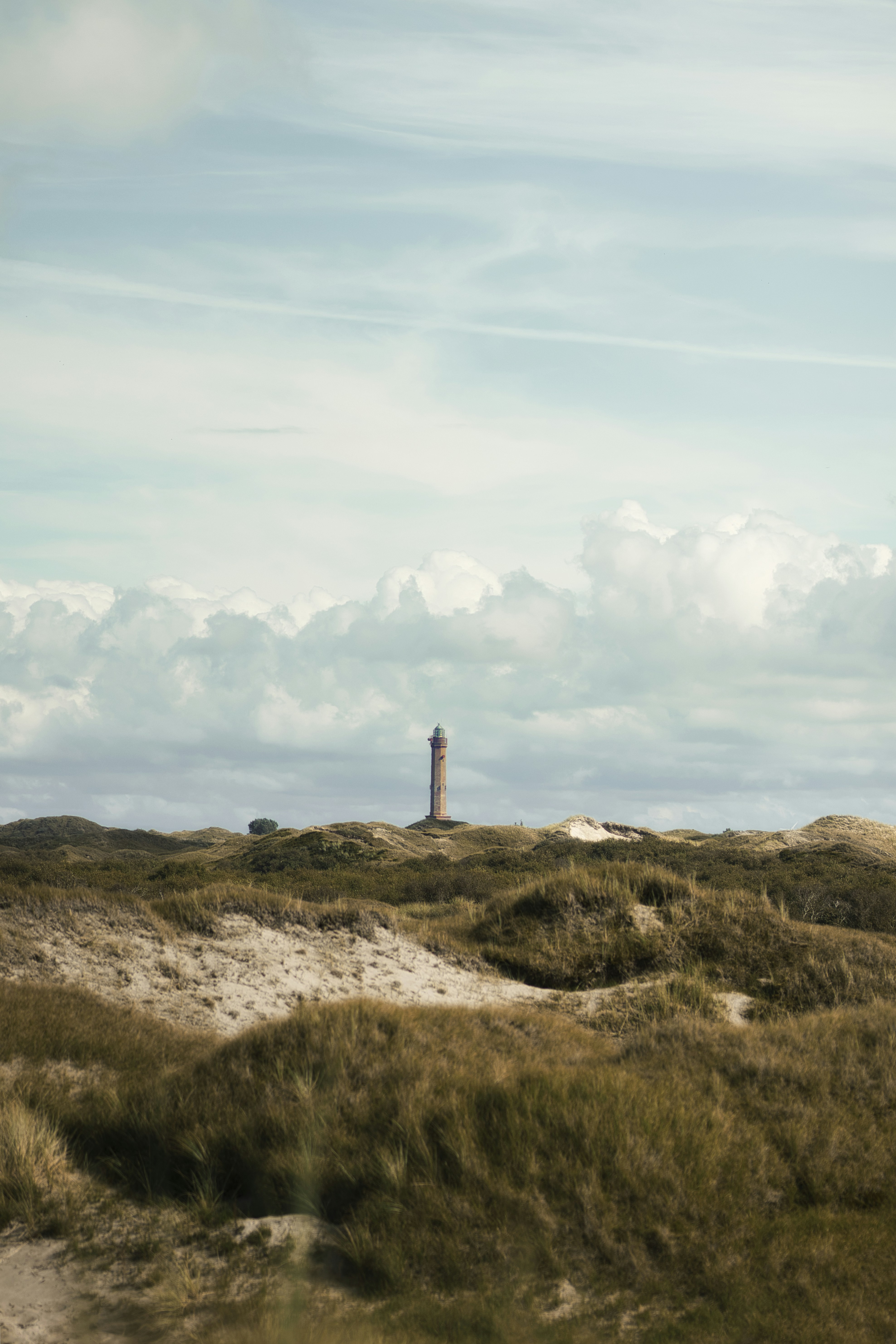 A tall lighthouse rising above grassy sand dunes under a cloudy blue sky