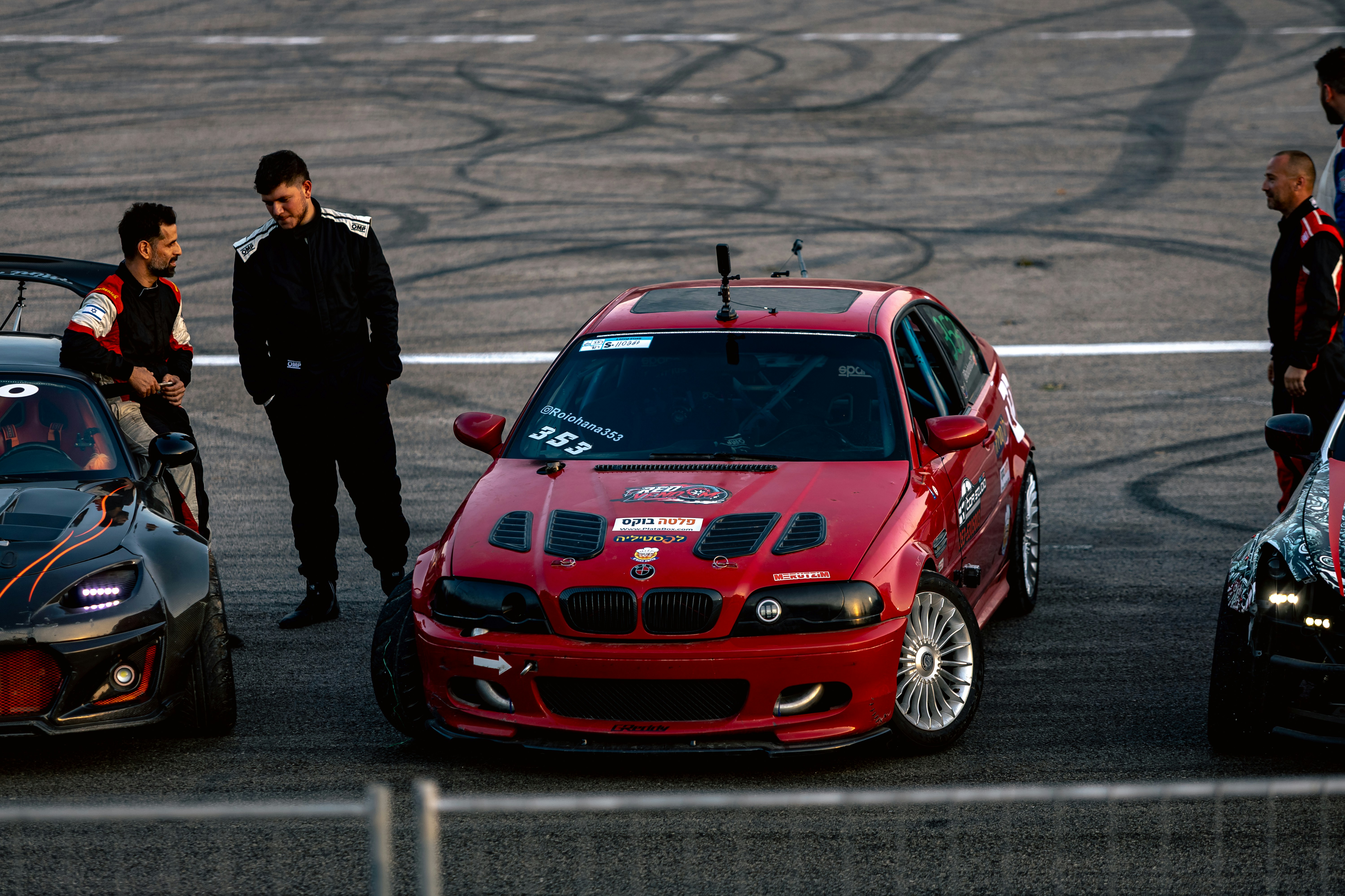 Three people in racing suits standing near modified drift cars on a track