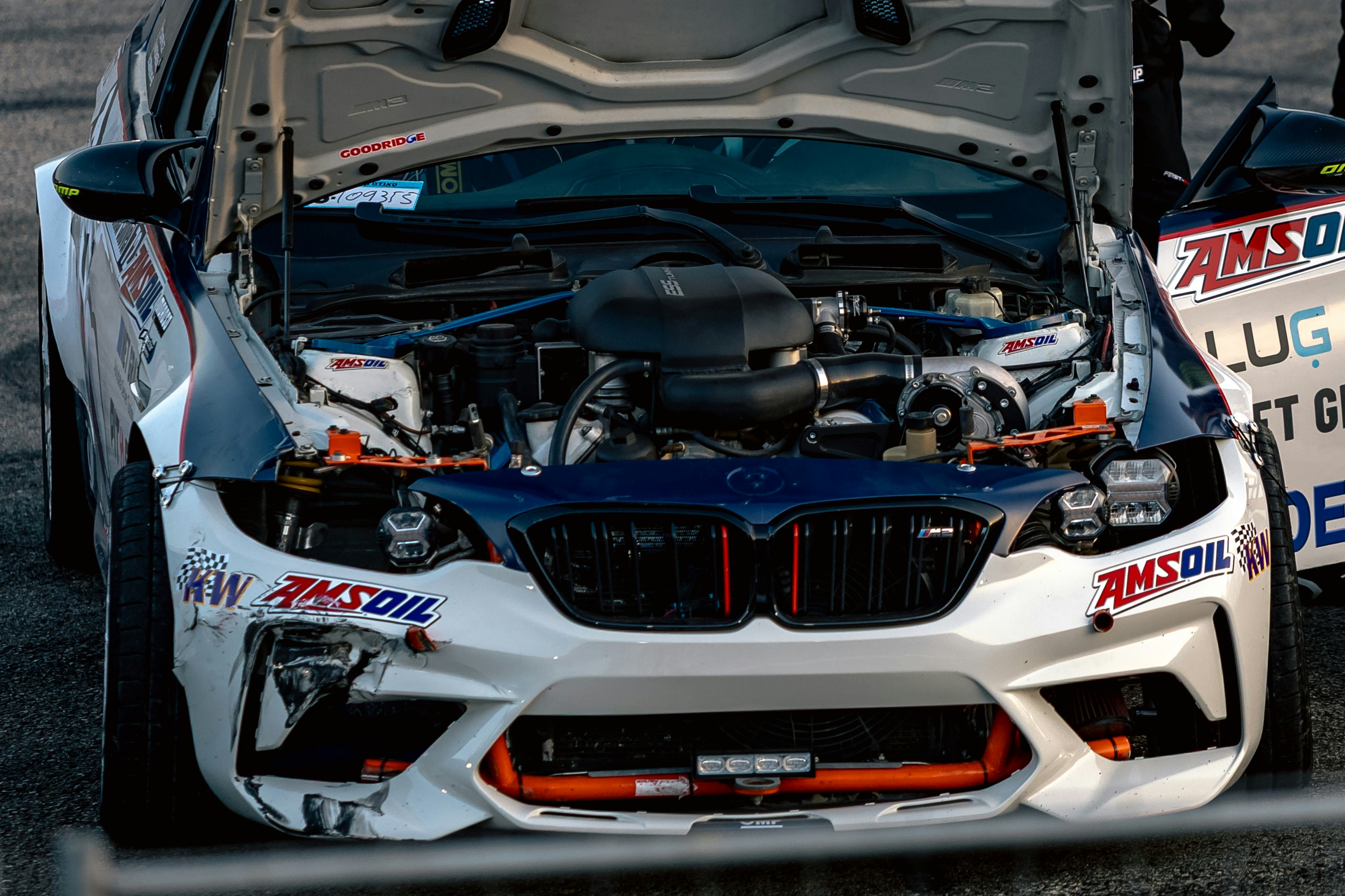 A white race car with an open hood showing the engine and damaged bumper