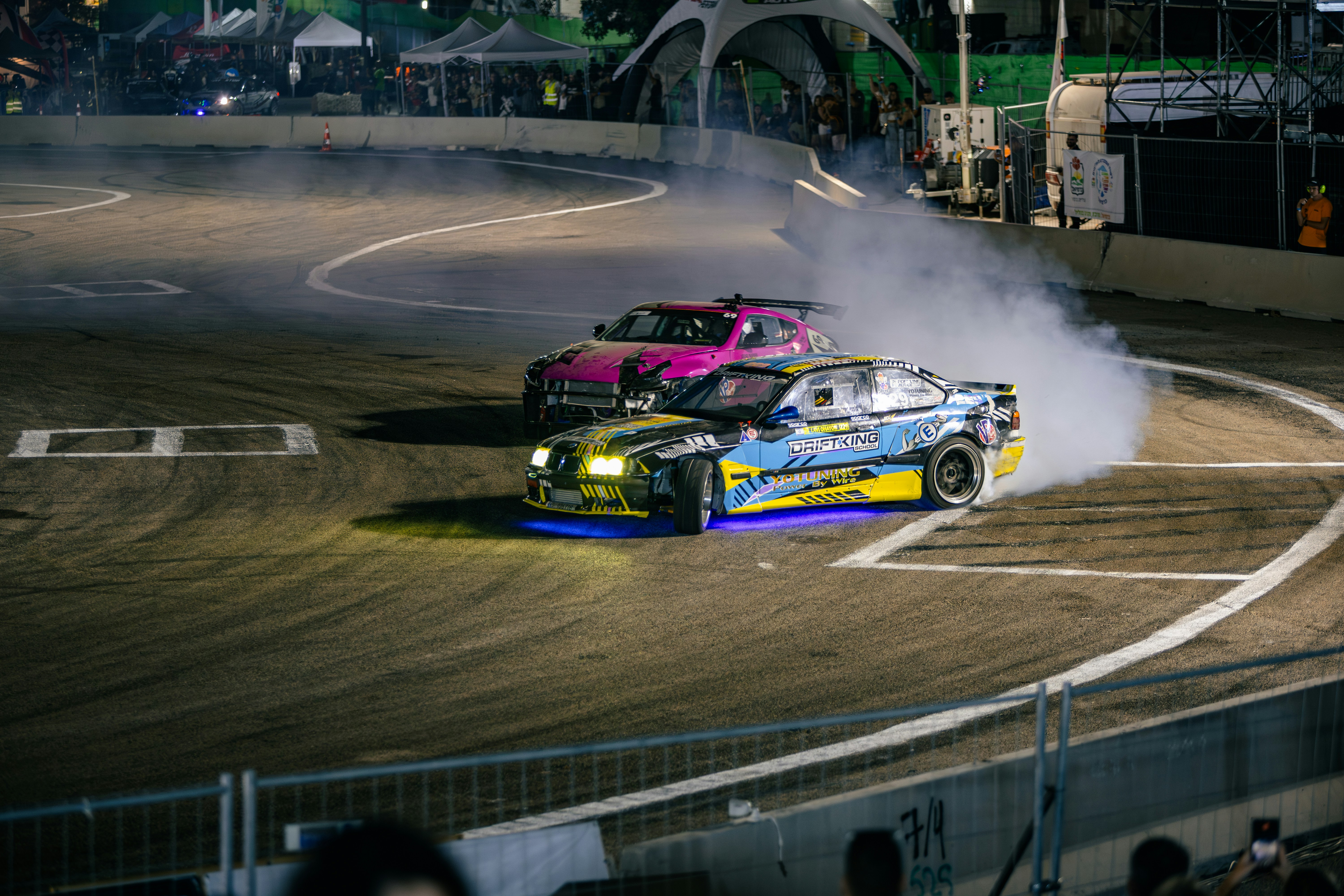 Two drift cars racing on a paved track at night with tire smoke