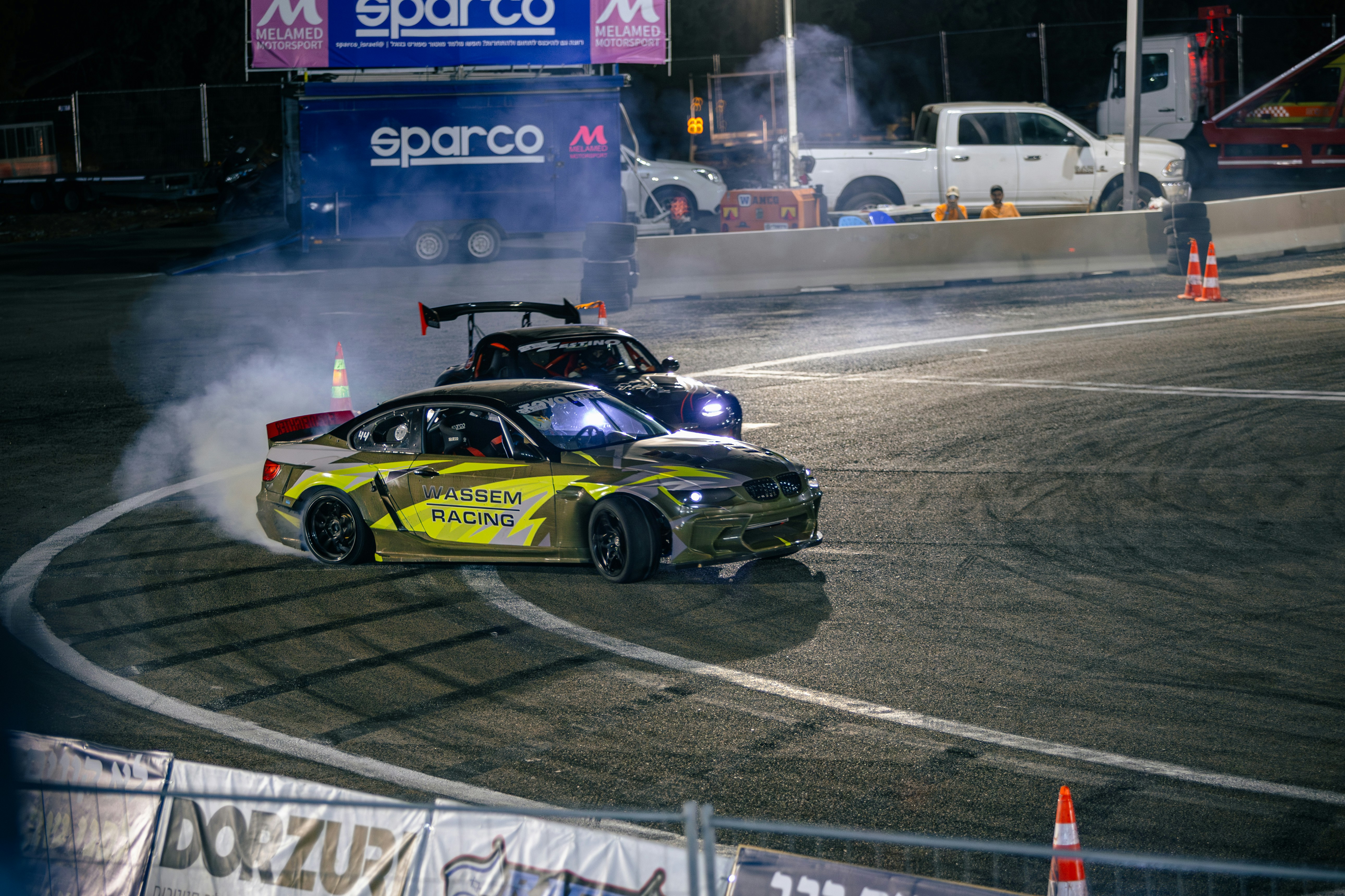 Two drift cars racing on an asphalt track at night with tire smoke