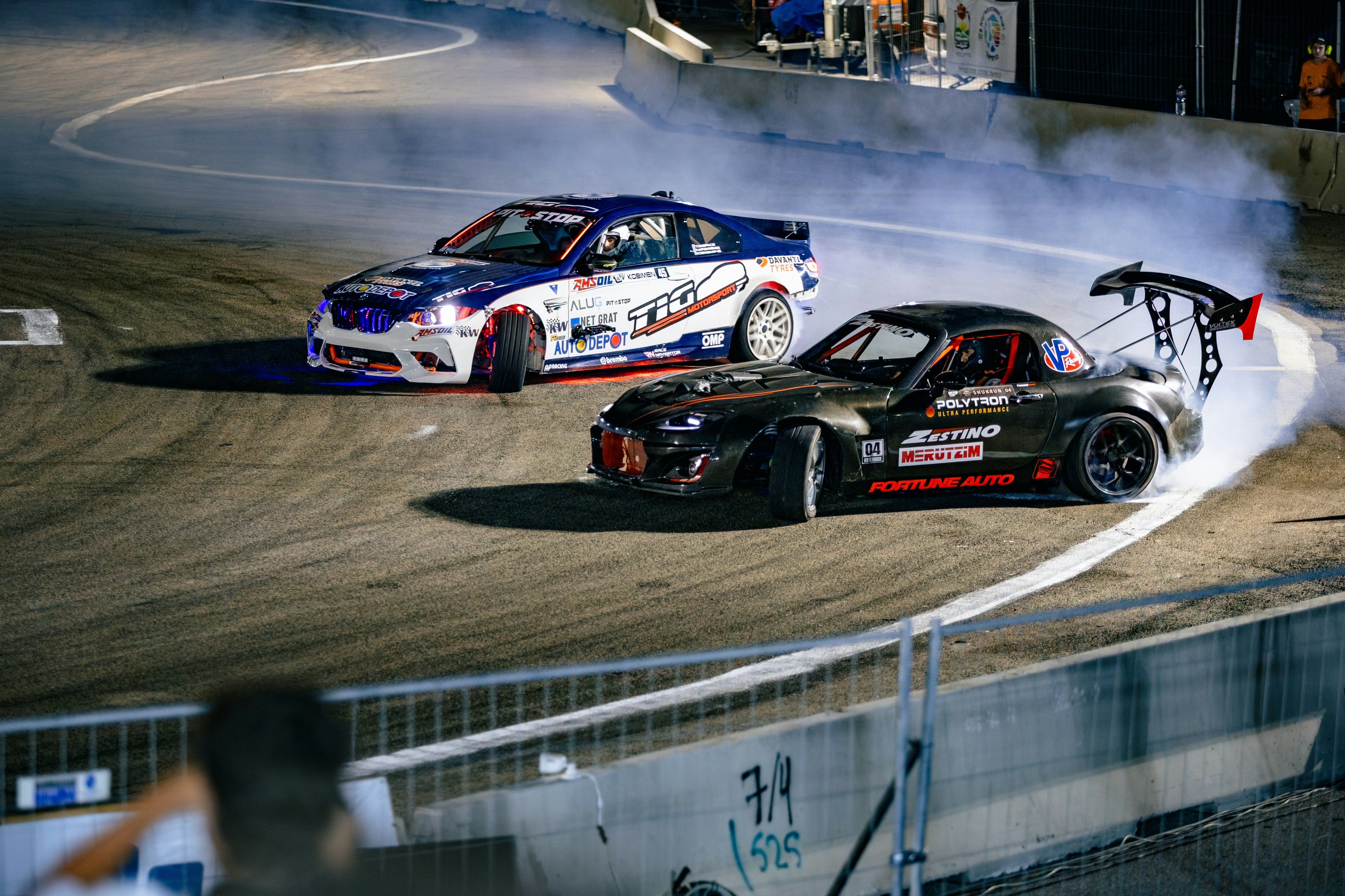 Two drift cars racing on an asphalt track with smoke from spinning tires