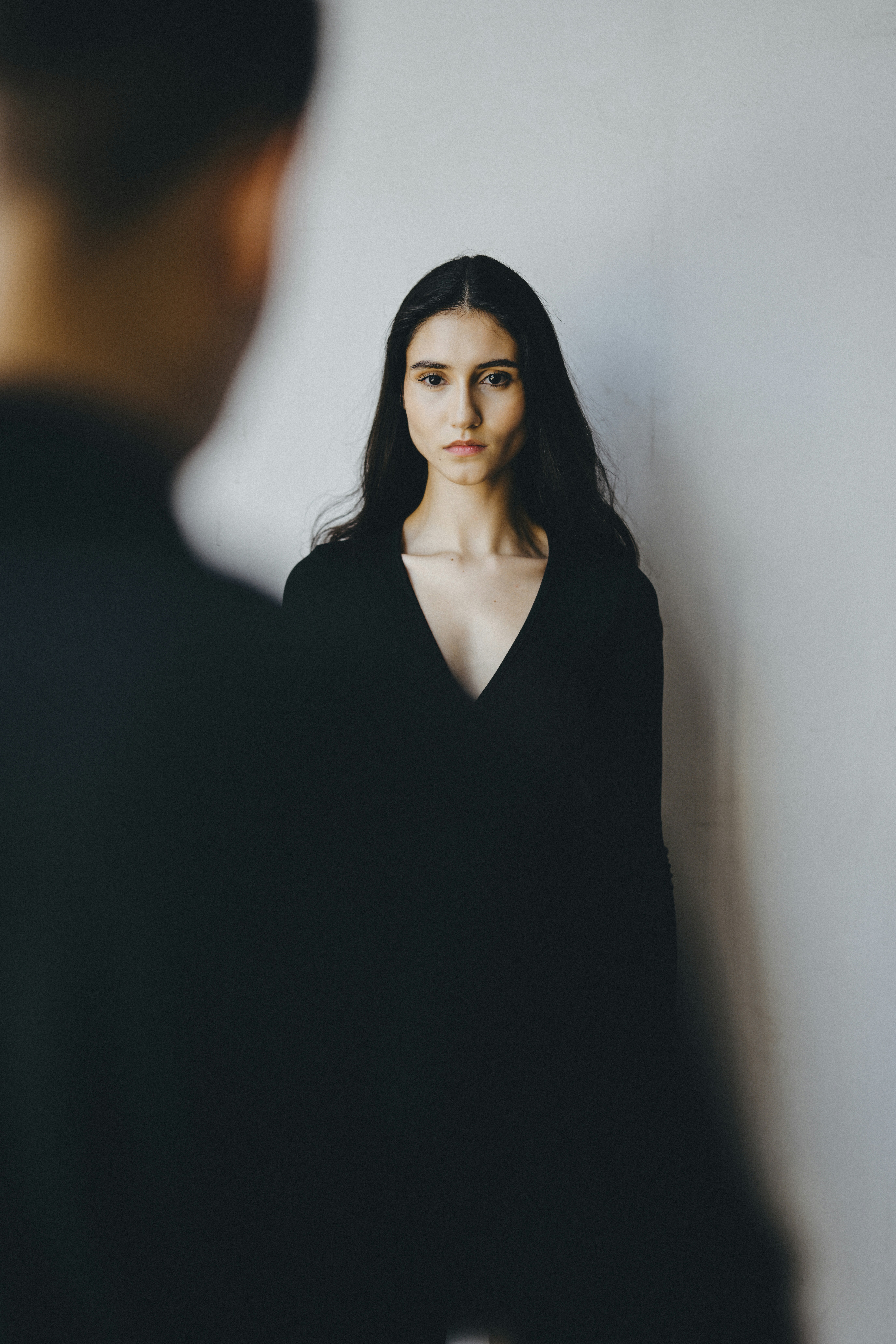 A woman with long dark hair in a black v-neck top against a wall