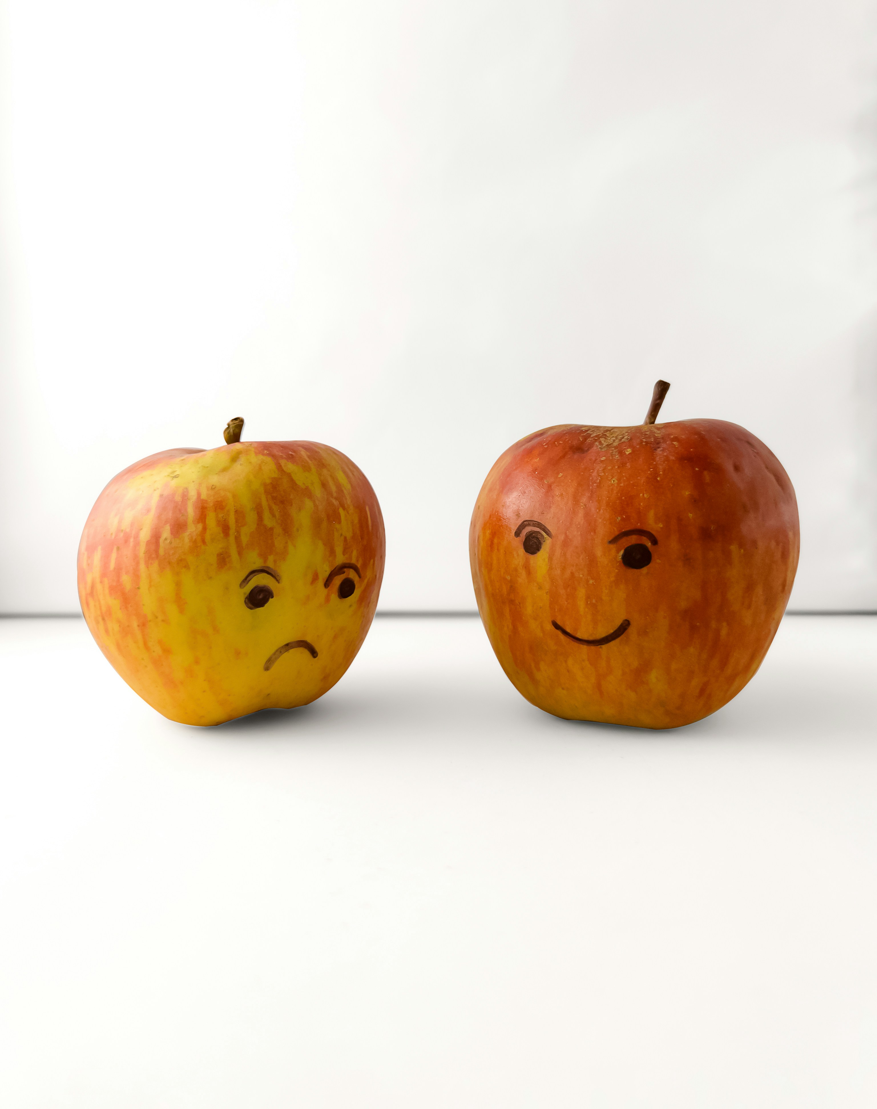 Two apples with drawn faces, one sad and one happy, on a white surface