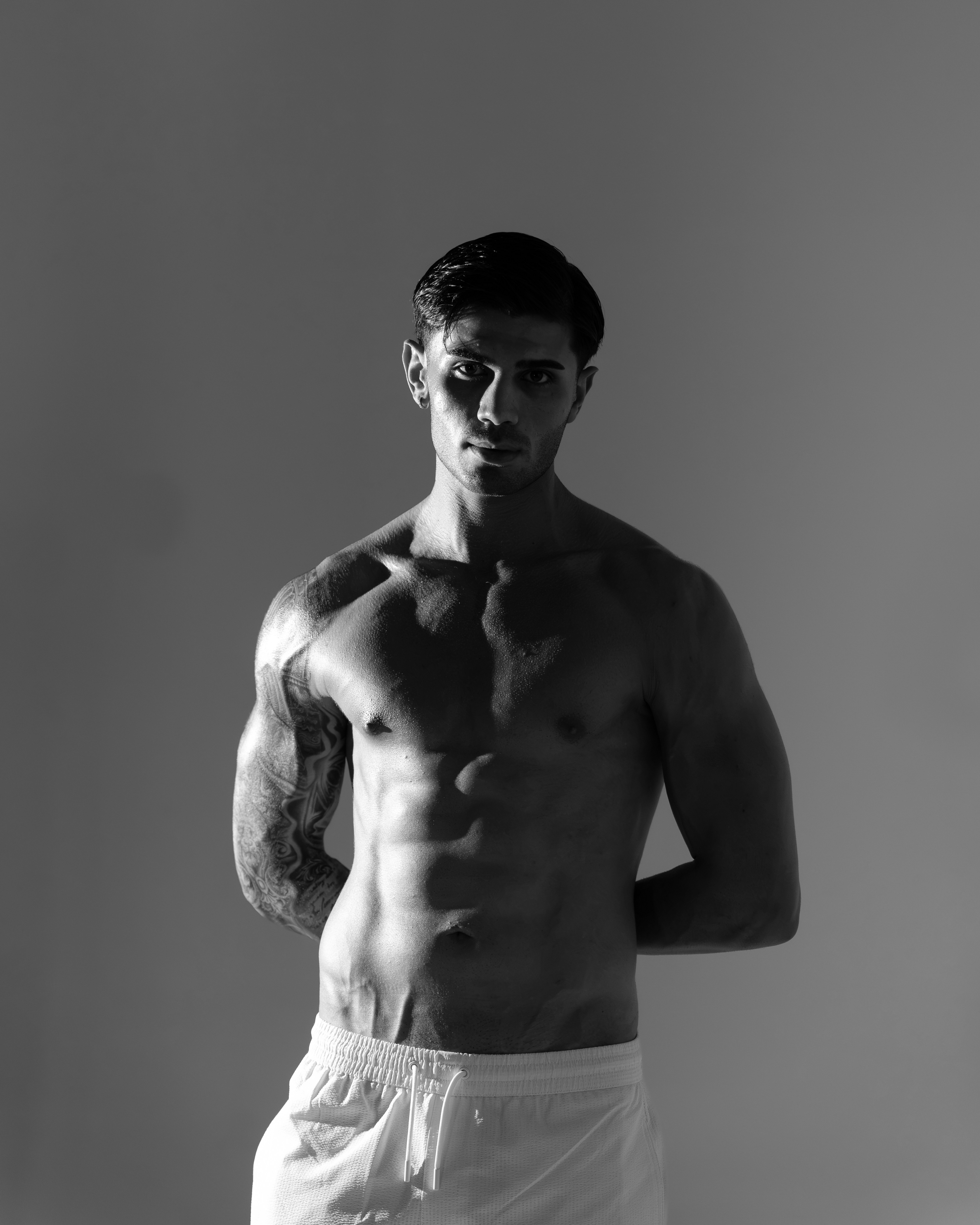 Um homem sem camisa com um braço tatuado, shorts brancos sobre um fundo cinza
