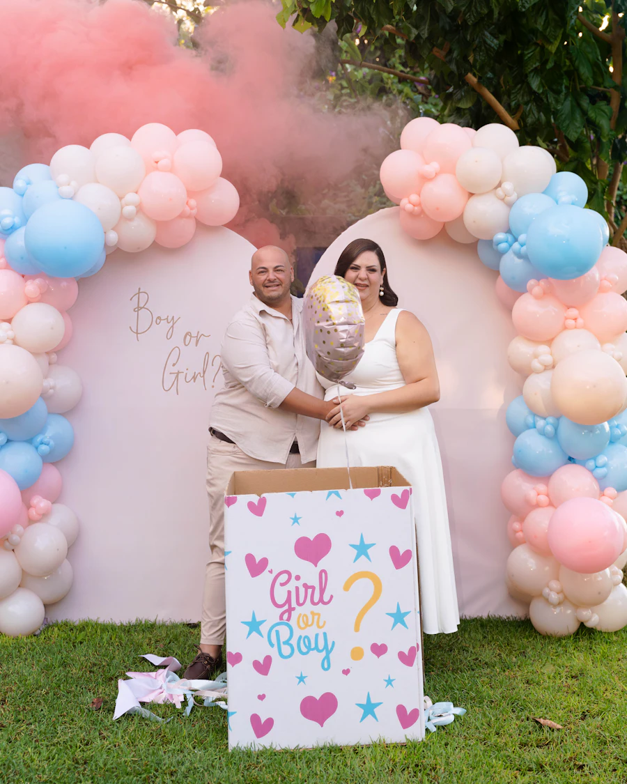 Couple at a gender reveal with pink smoke behind a pink and blue balloon arch