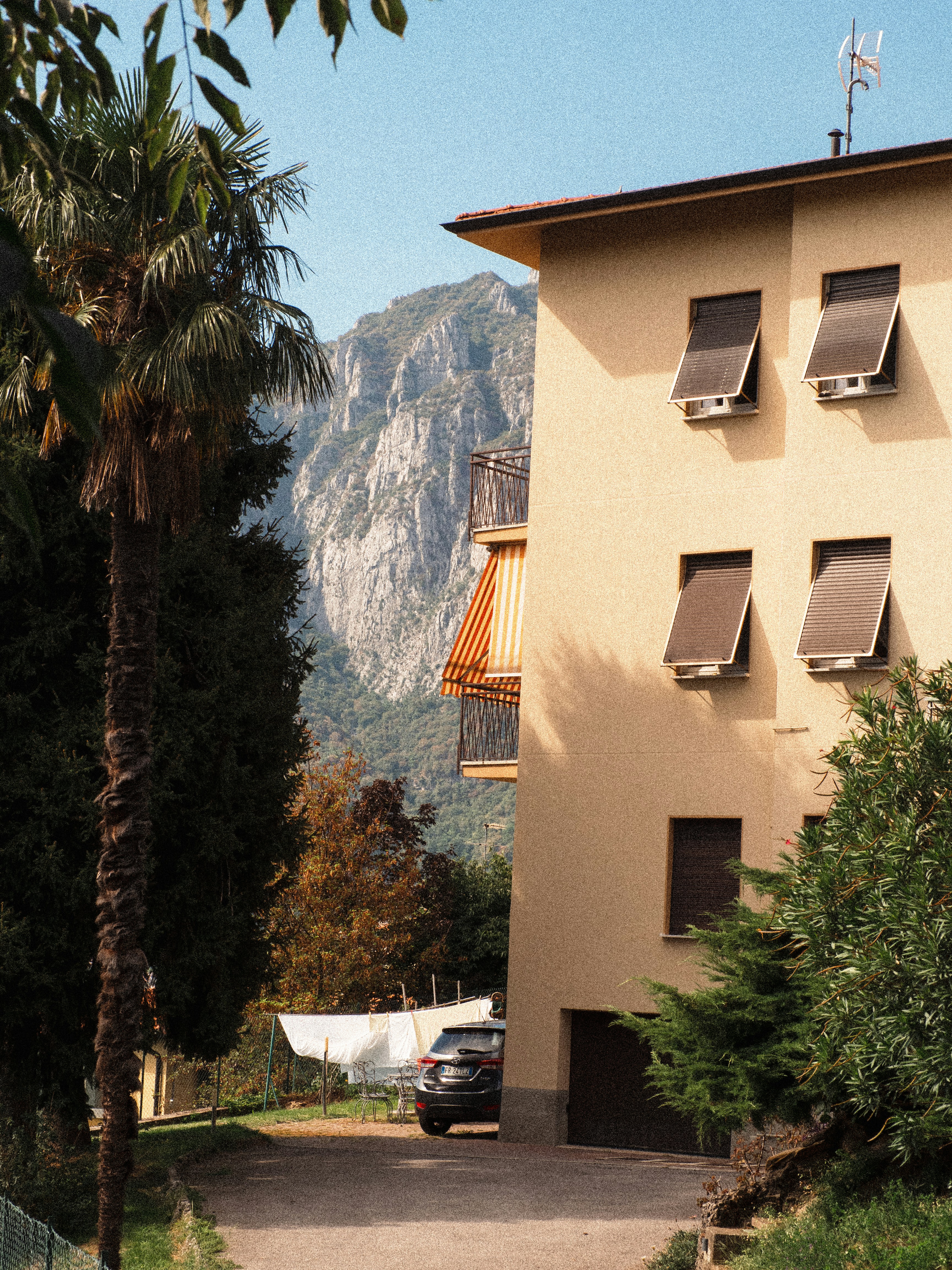 A beige apartment building with brown shutters beside a palm tree and mountain
