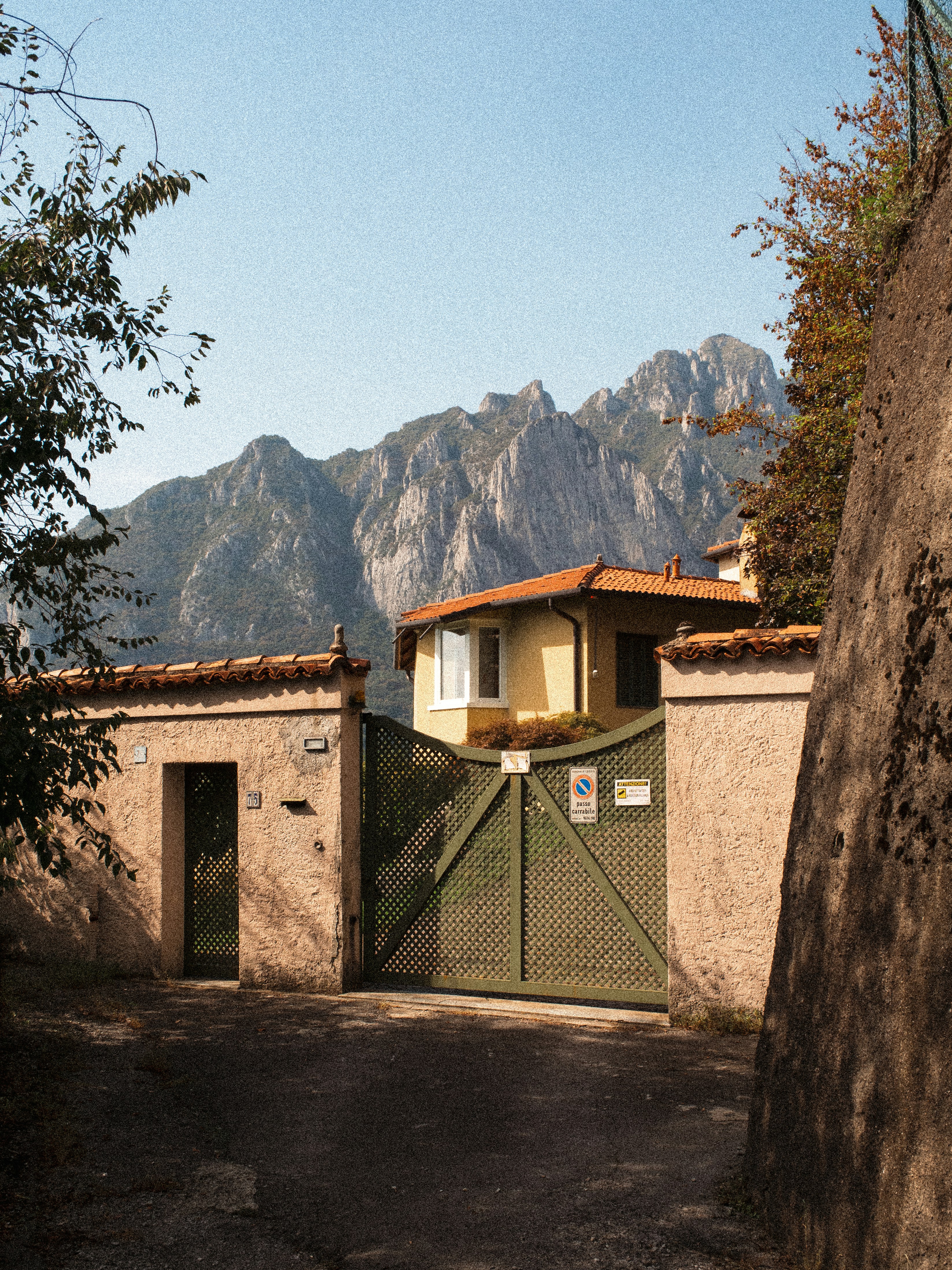 A yellow house behind a green gate and stone wall with mountains behind
