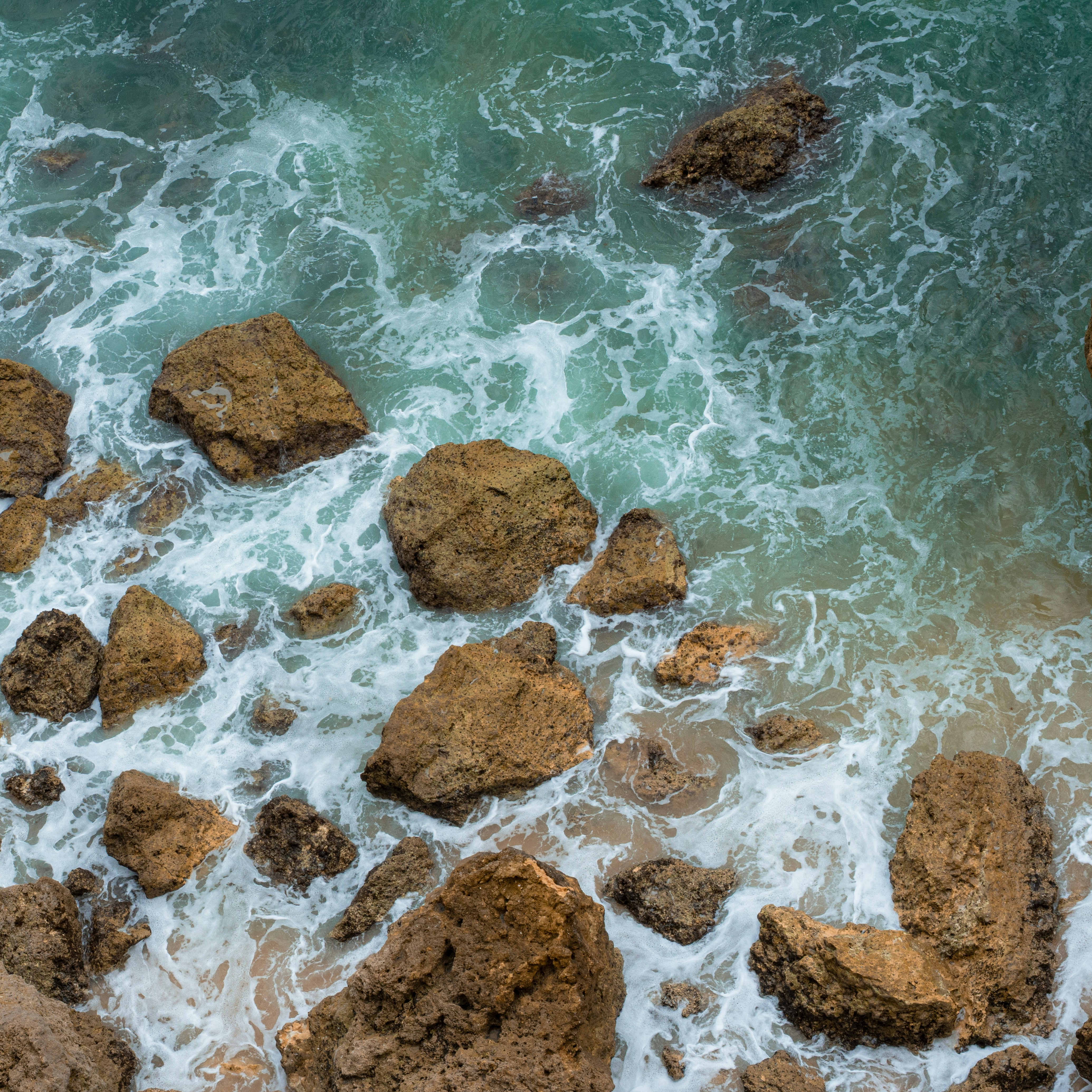 Aerial view of brown rocks surrounded by white sea foam and turquoise water