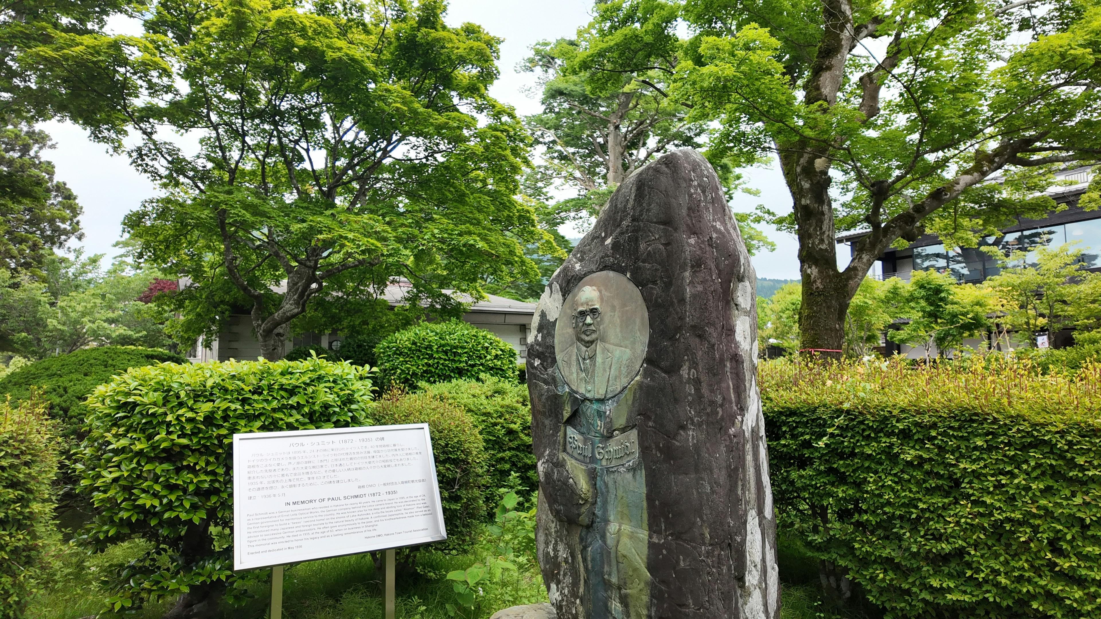 A stone monument with a circular portrait inset stands beside a white informational plaque, surrounded by trimmed hedges and lush trees in a park-like setting.