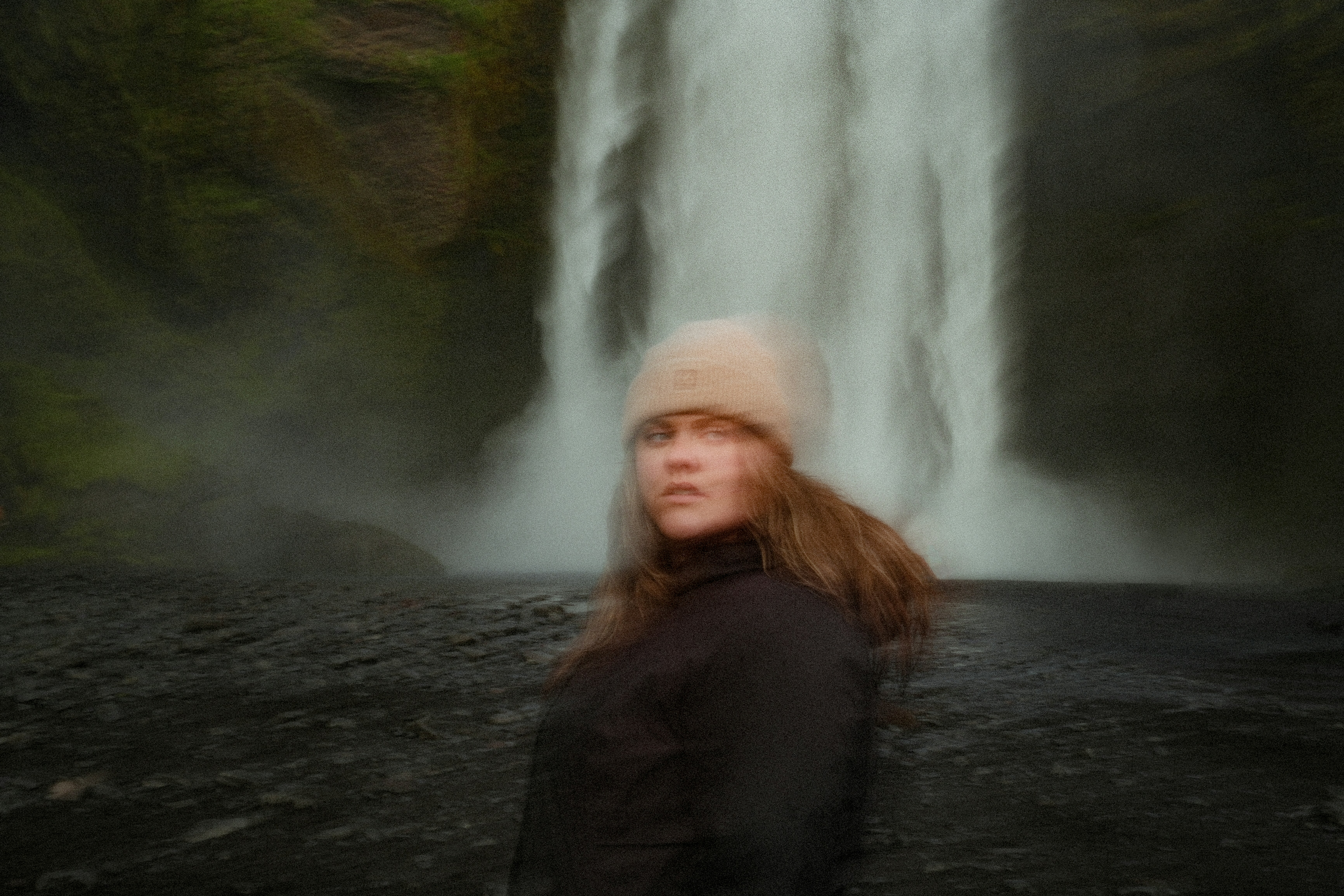 Une personne en bonnet beige et veste noire devant une cascade en Islande