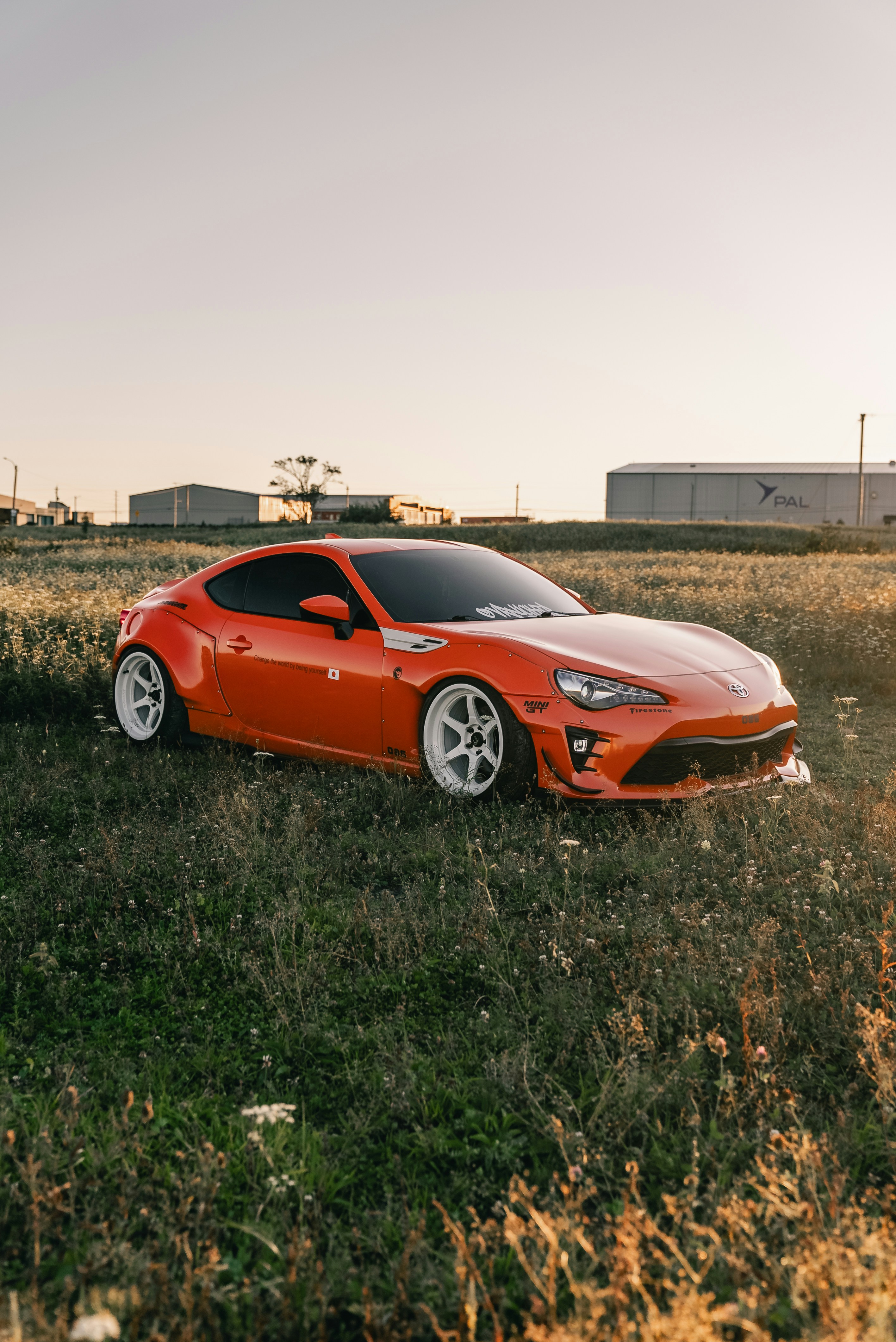 An orange sports car with white wheels parked in a grassy field at golden hour