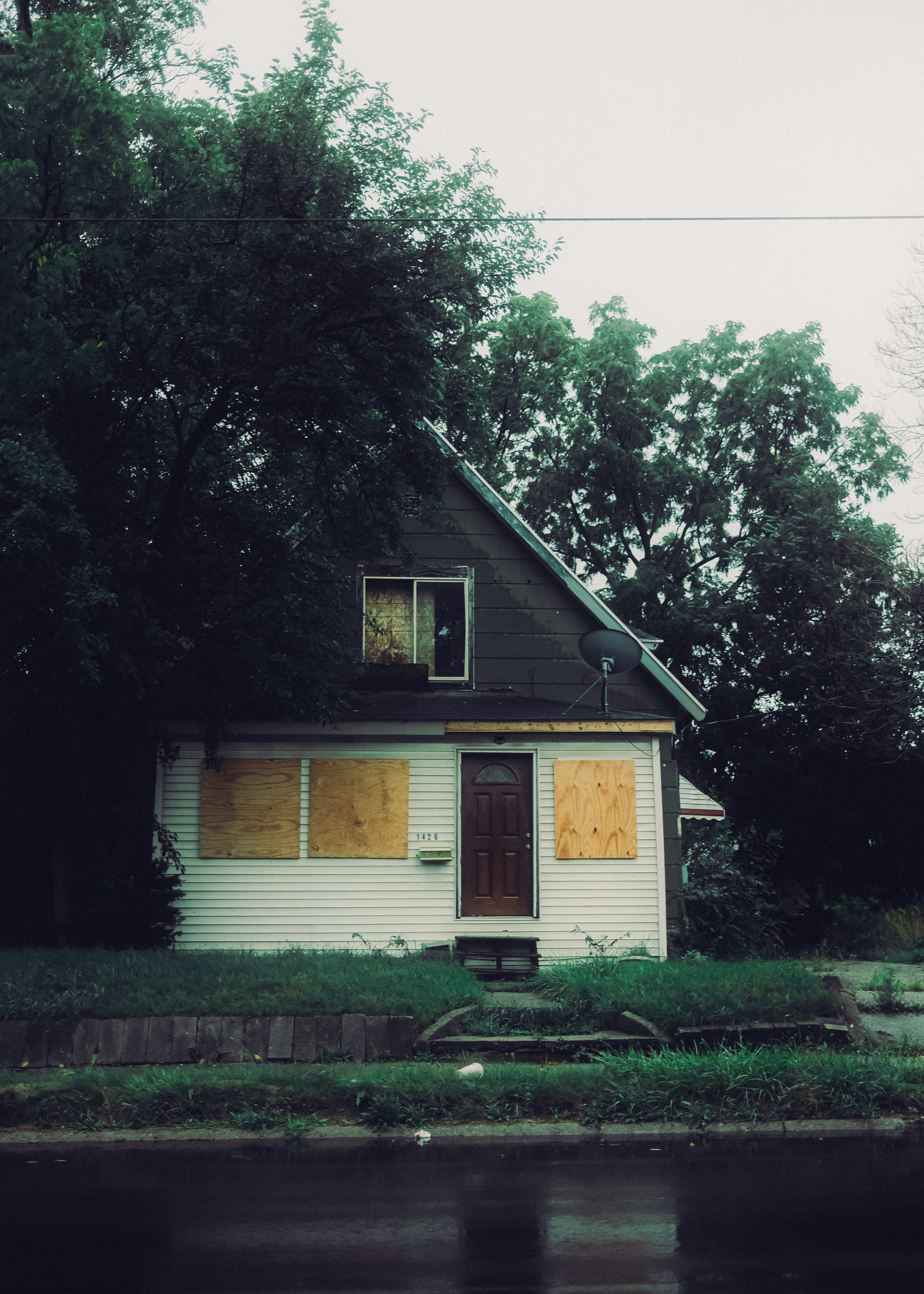 A house with boarded windows and a brown door behind large green trees