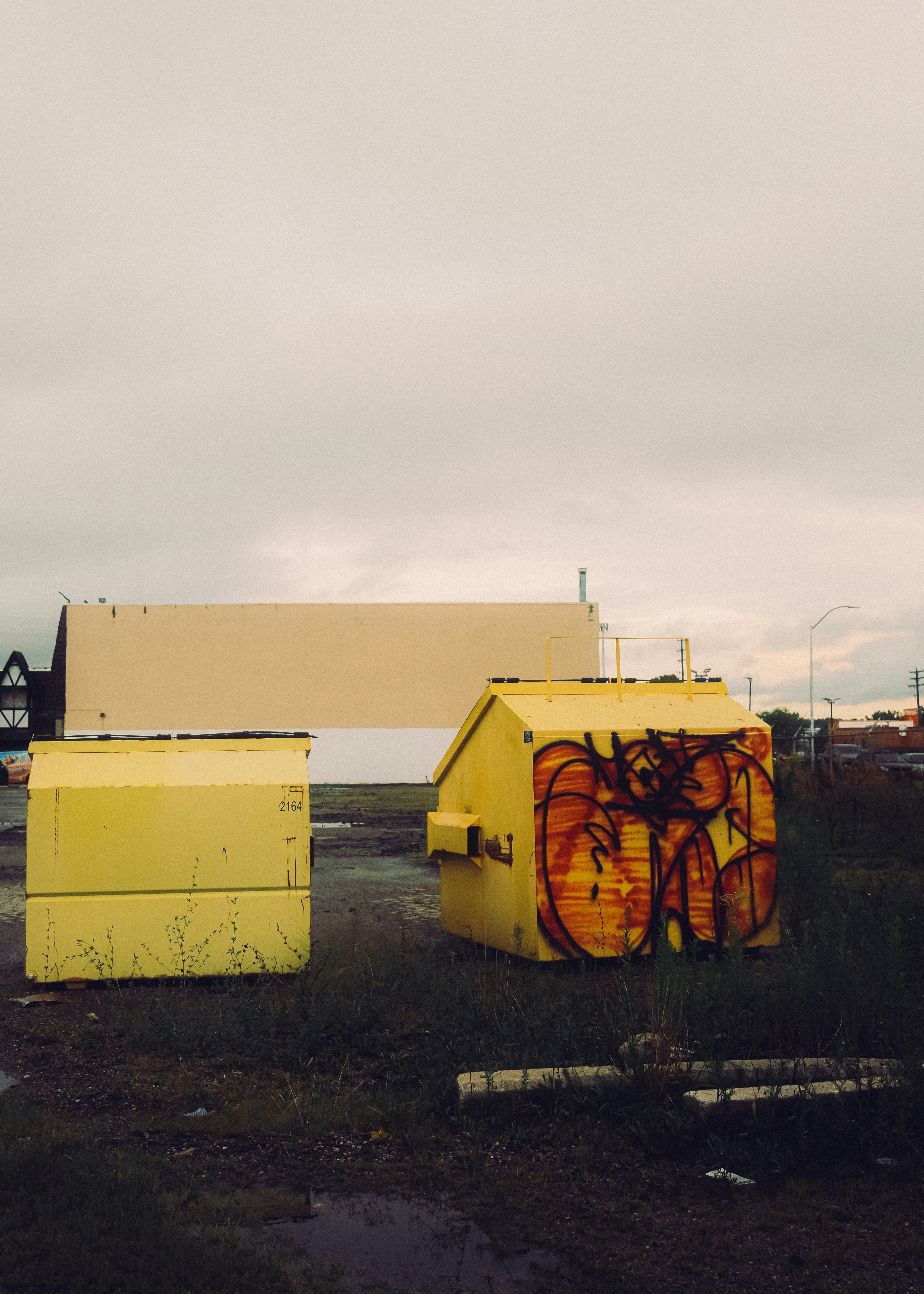 Two yellow dumpsters on a dirt lot with a large beige wall behind