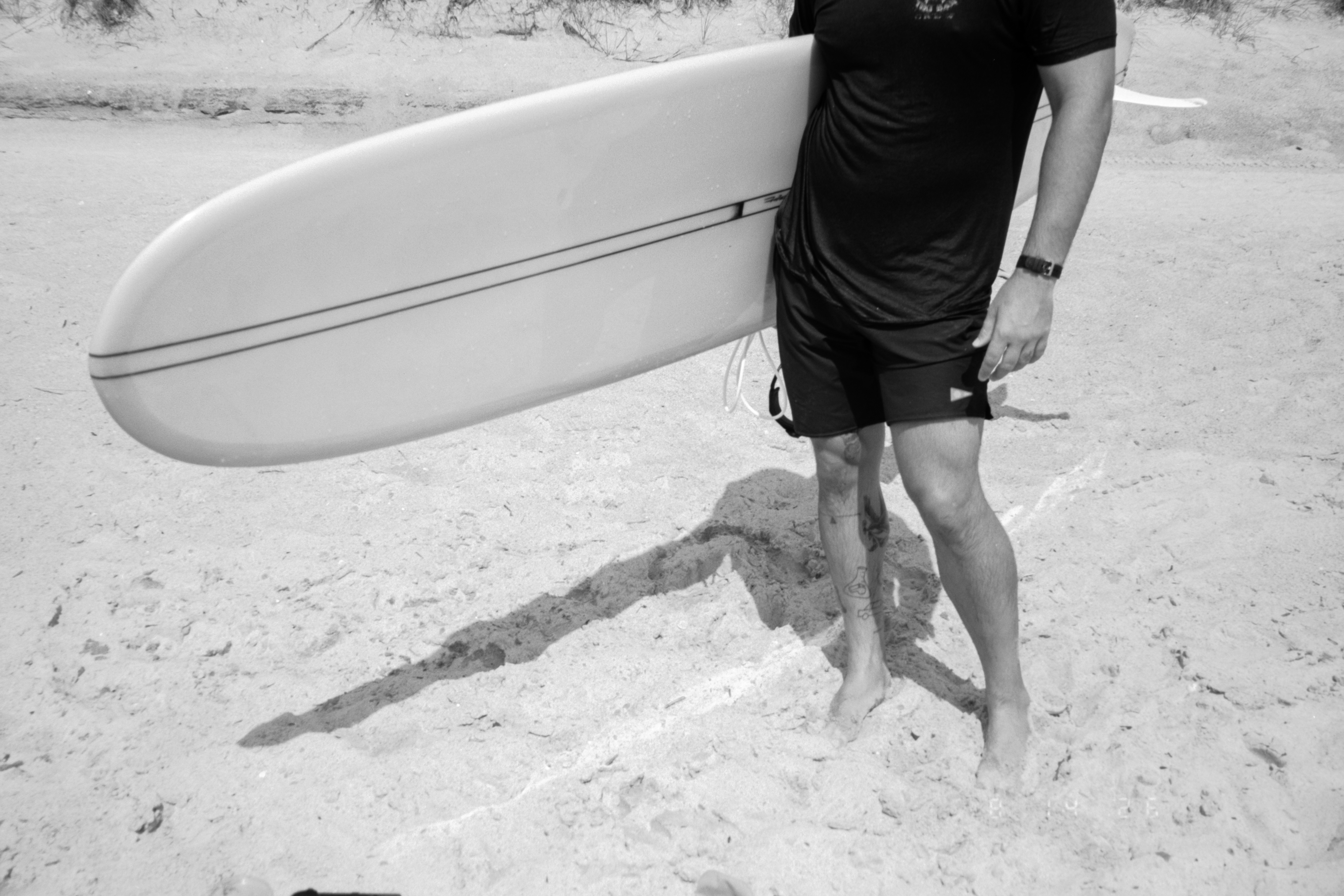 Una persona con pantalones cortos negros y camiseta sosteniendo una tabla de surf sobre la arena