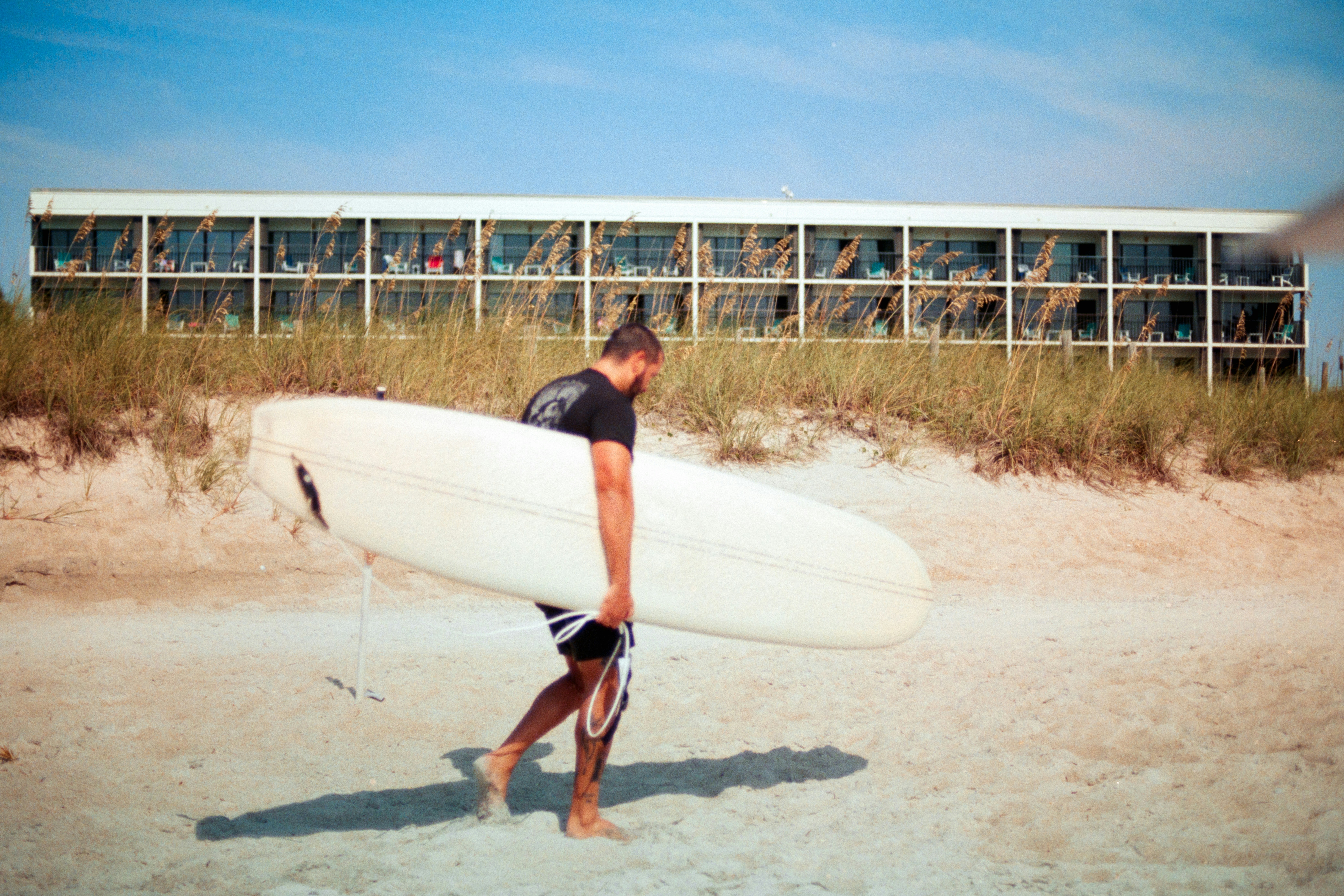 Un hombre que lleva una tabla de surf blanca a través de una playa de arena cerca de un hotel