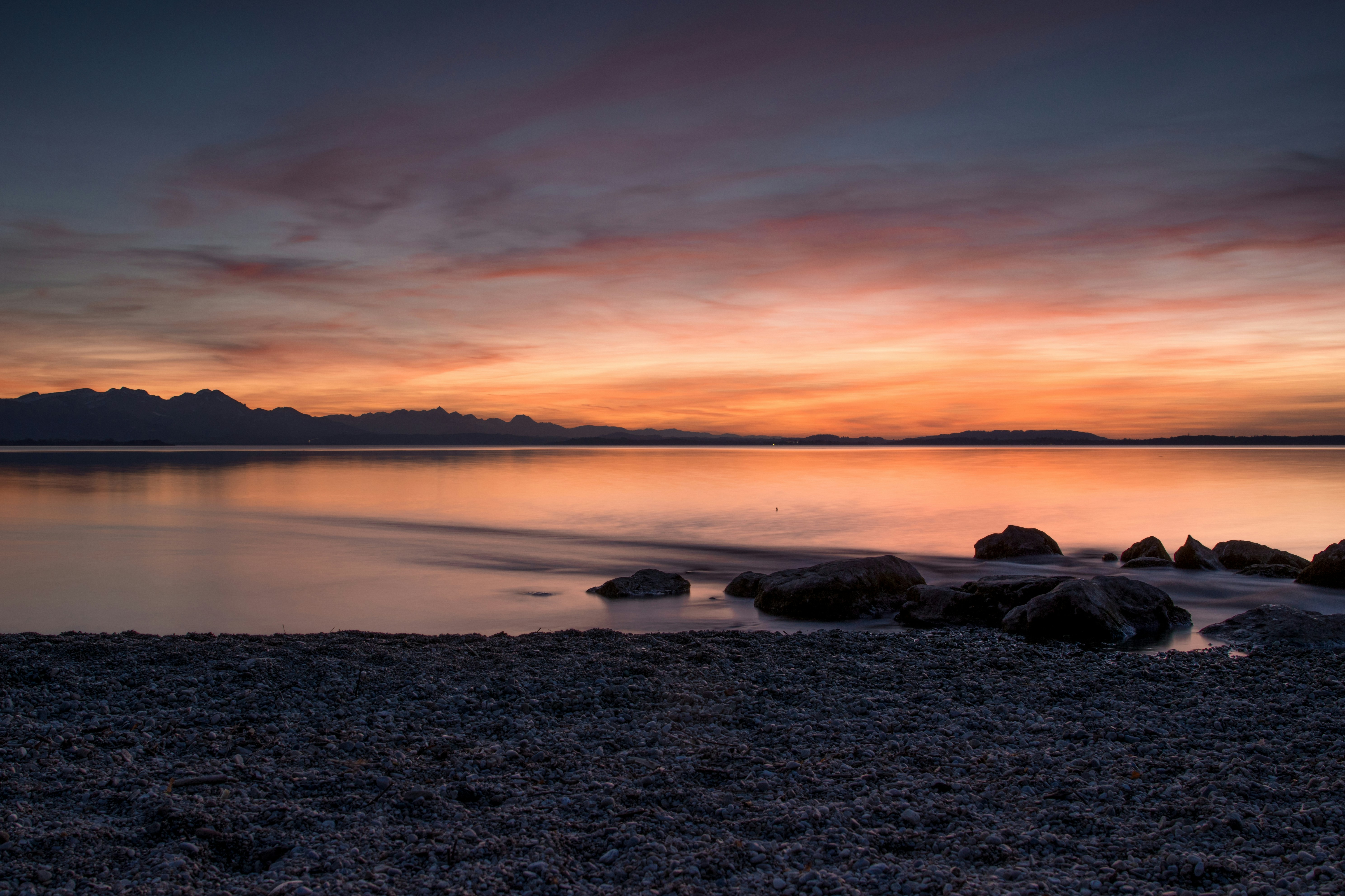 Ein ruhiger See mit dunklen Felsen an einem kieseligen Ufer unter einem warmen, orangefarbenen Himmel