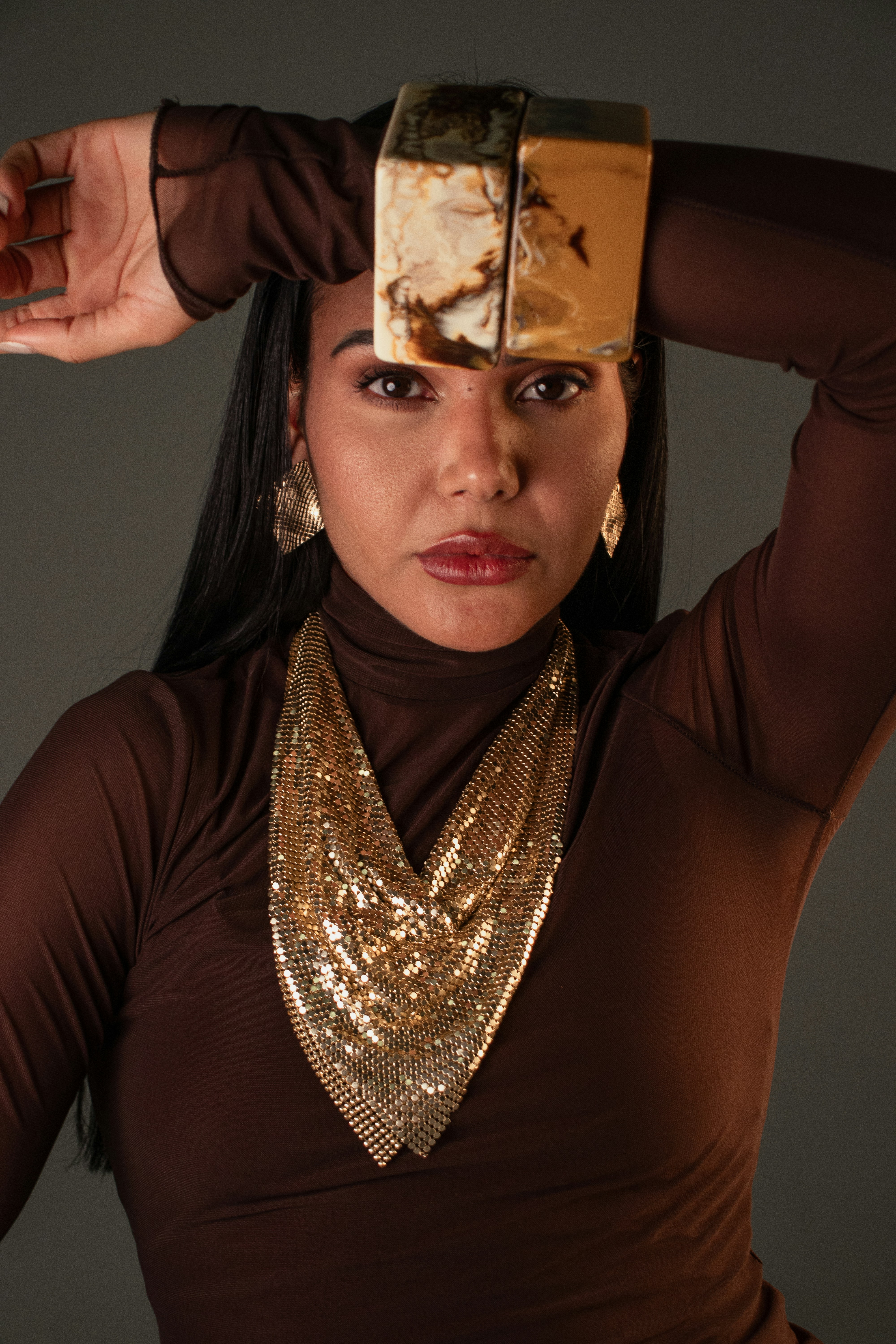 A woman in a brown turtleneck wearing gold chainmail jewelry and marble bracelets