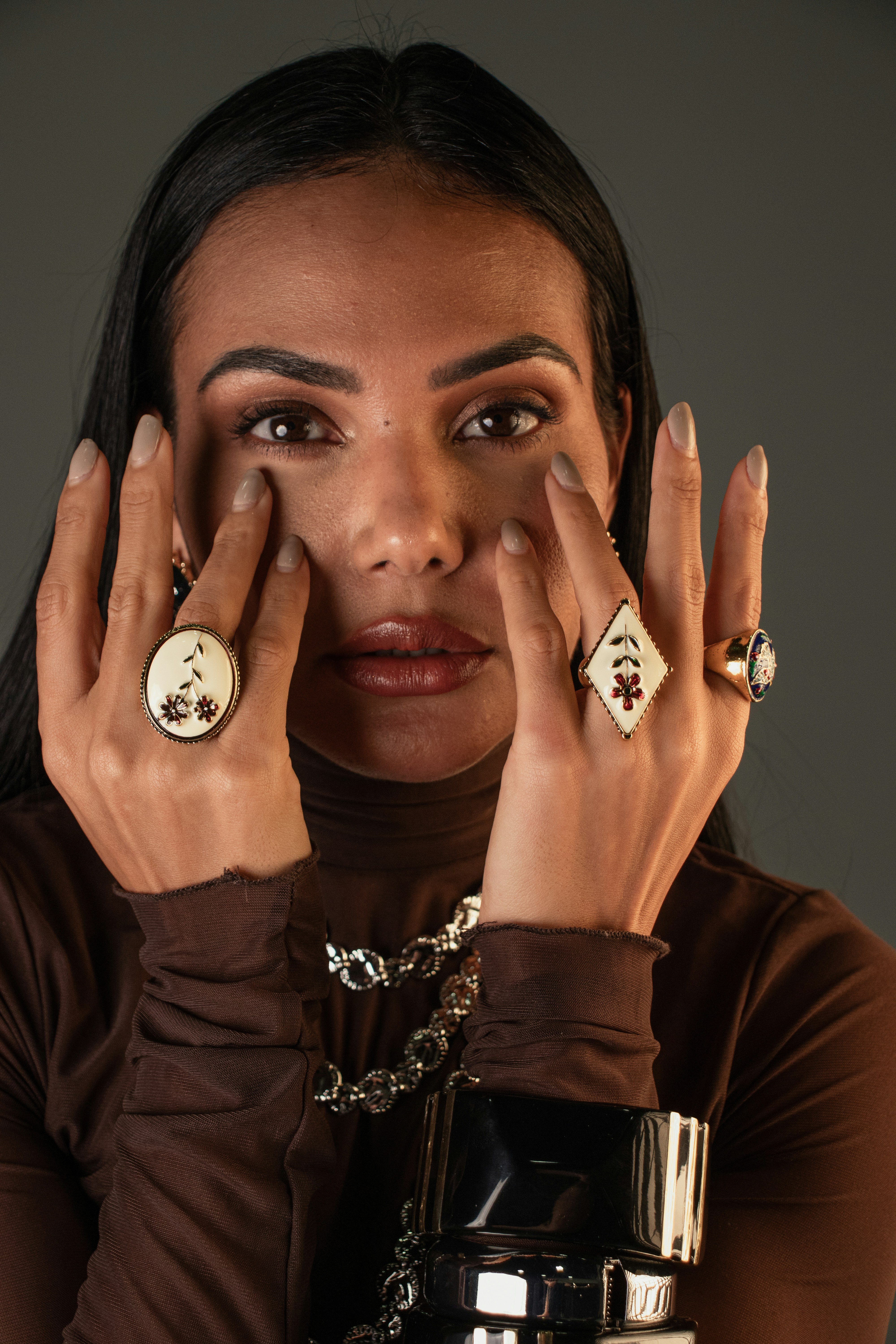 A woman wearing floral rings and a brown turtleneck top against a grey background