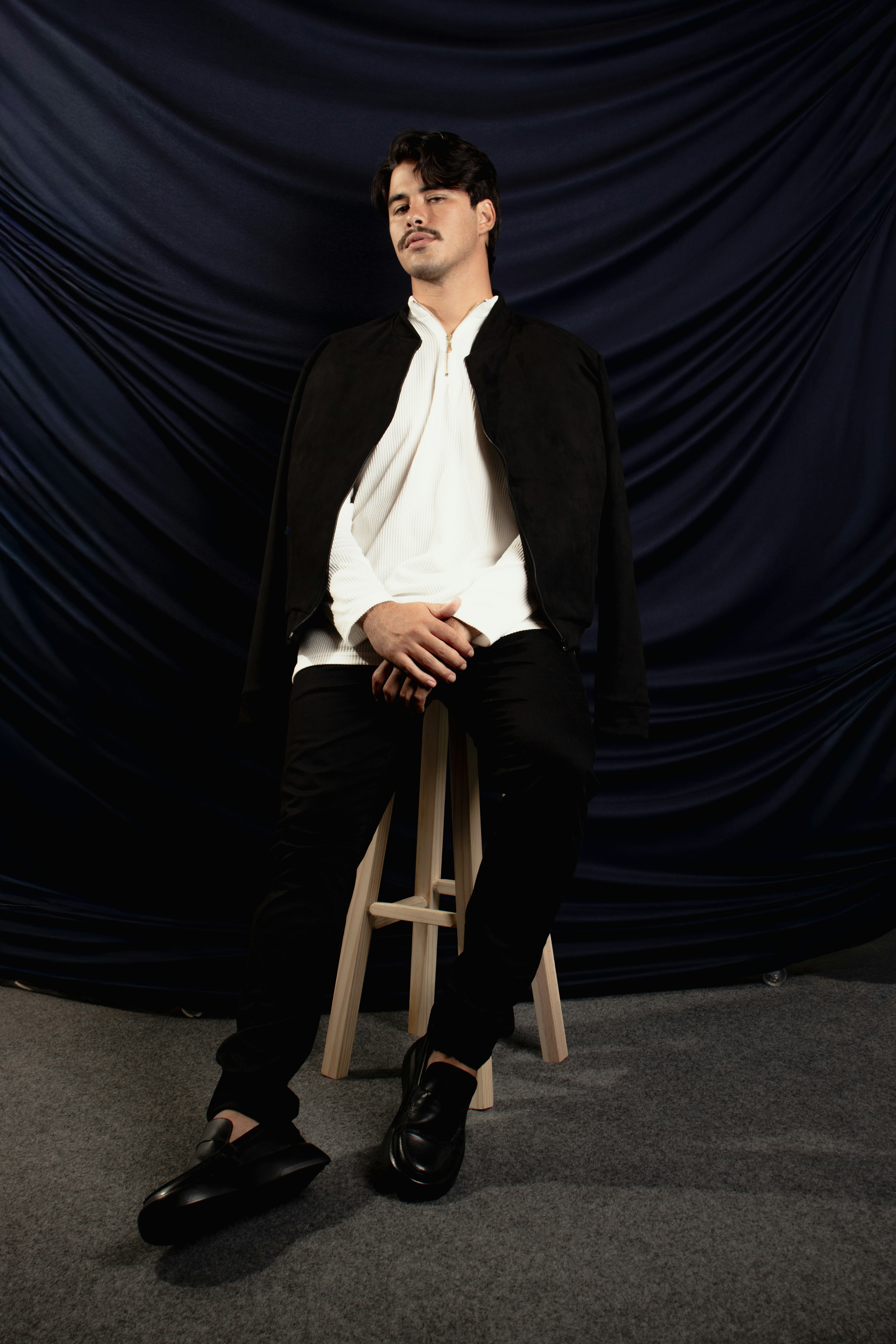 A man with a mustache in a white shirt and black jacket on a stool