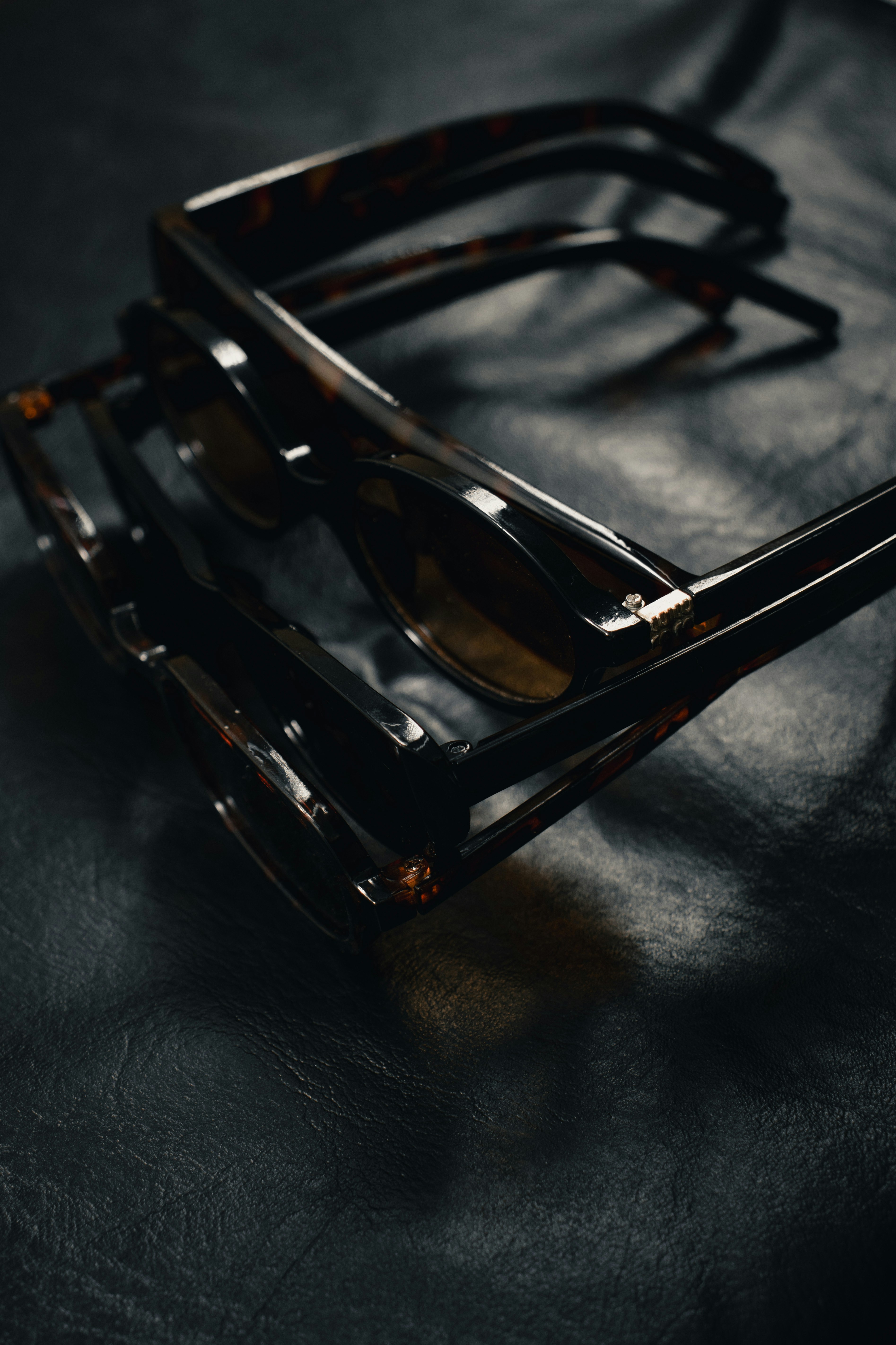 Two pairs of tortoiseshell sunglasses stacked on a dark textured leather surface
