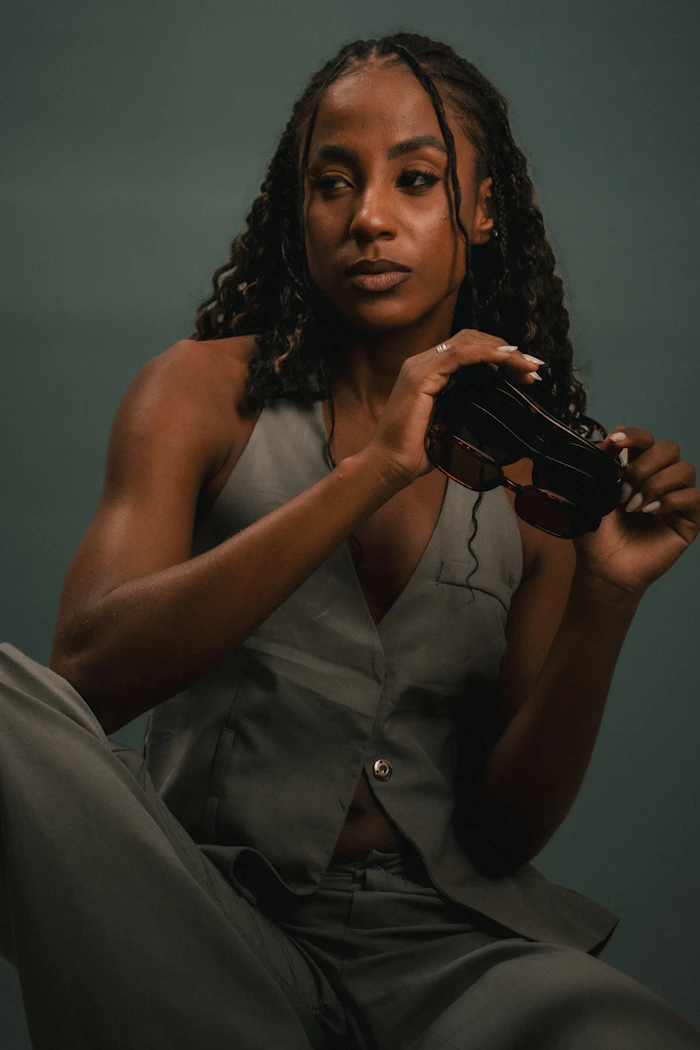 A woman in a grey tailored vest holding a pair of sunglasses, photographed against a dark green wall.