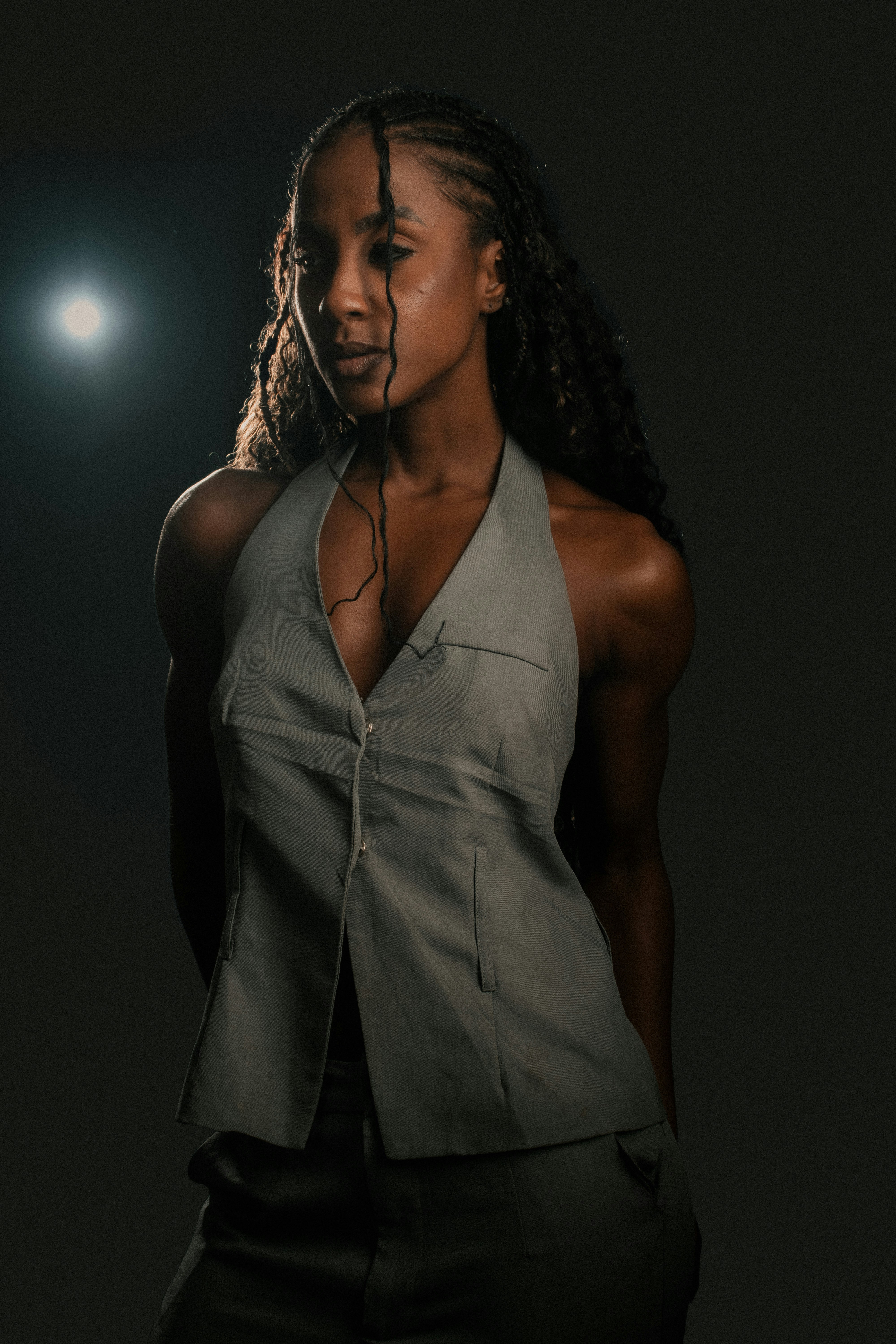A woman with braided hair wearing a grey vest against a dark background