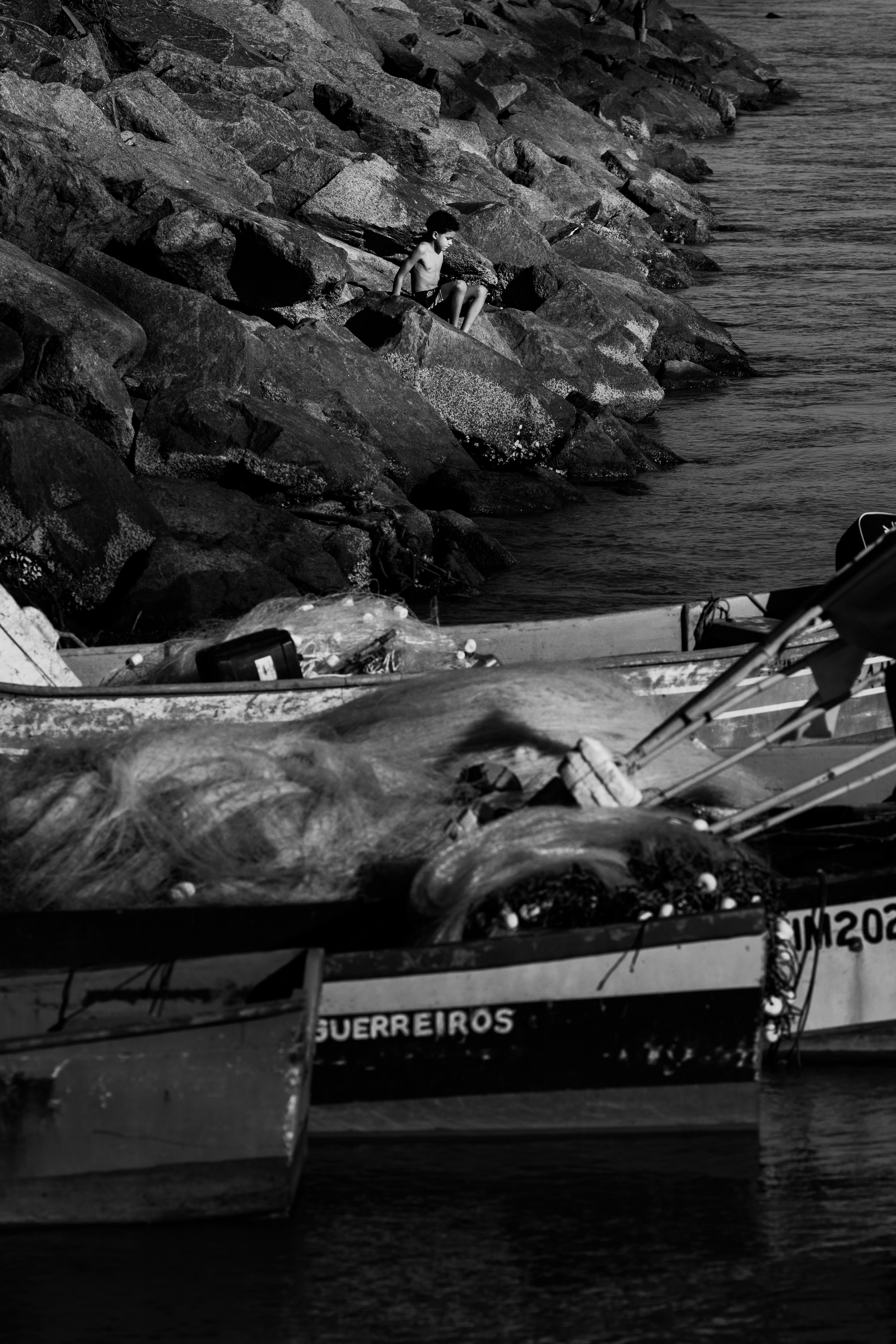 Un niño en una orilla rocosa cerca de barcos pesqueros con redes