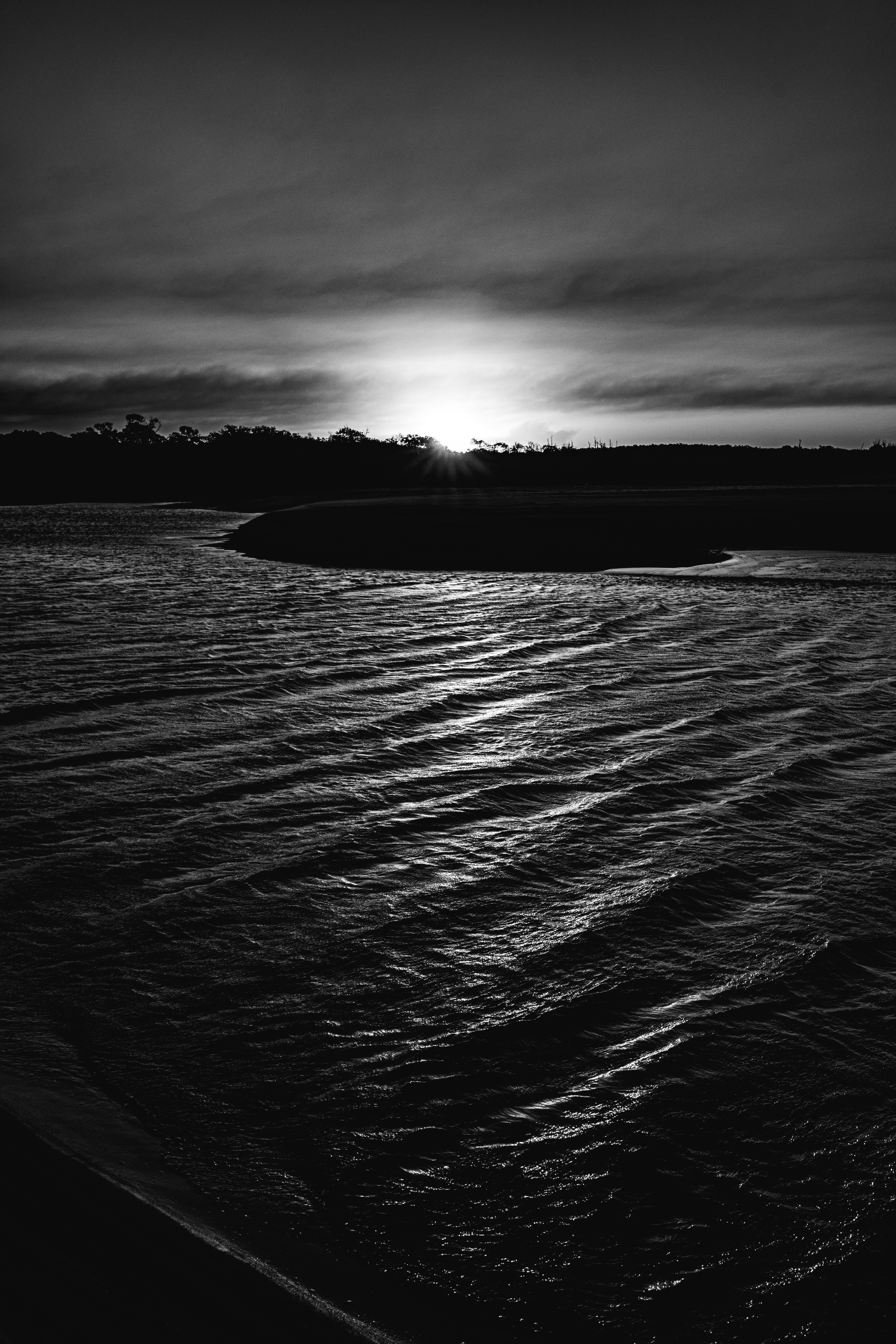 Sol bajo sobre agua ondulante y una orilla oscura en blanco y negro