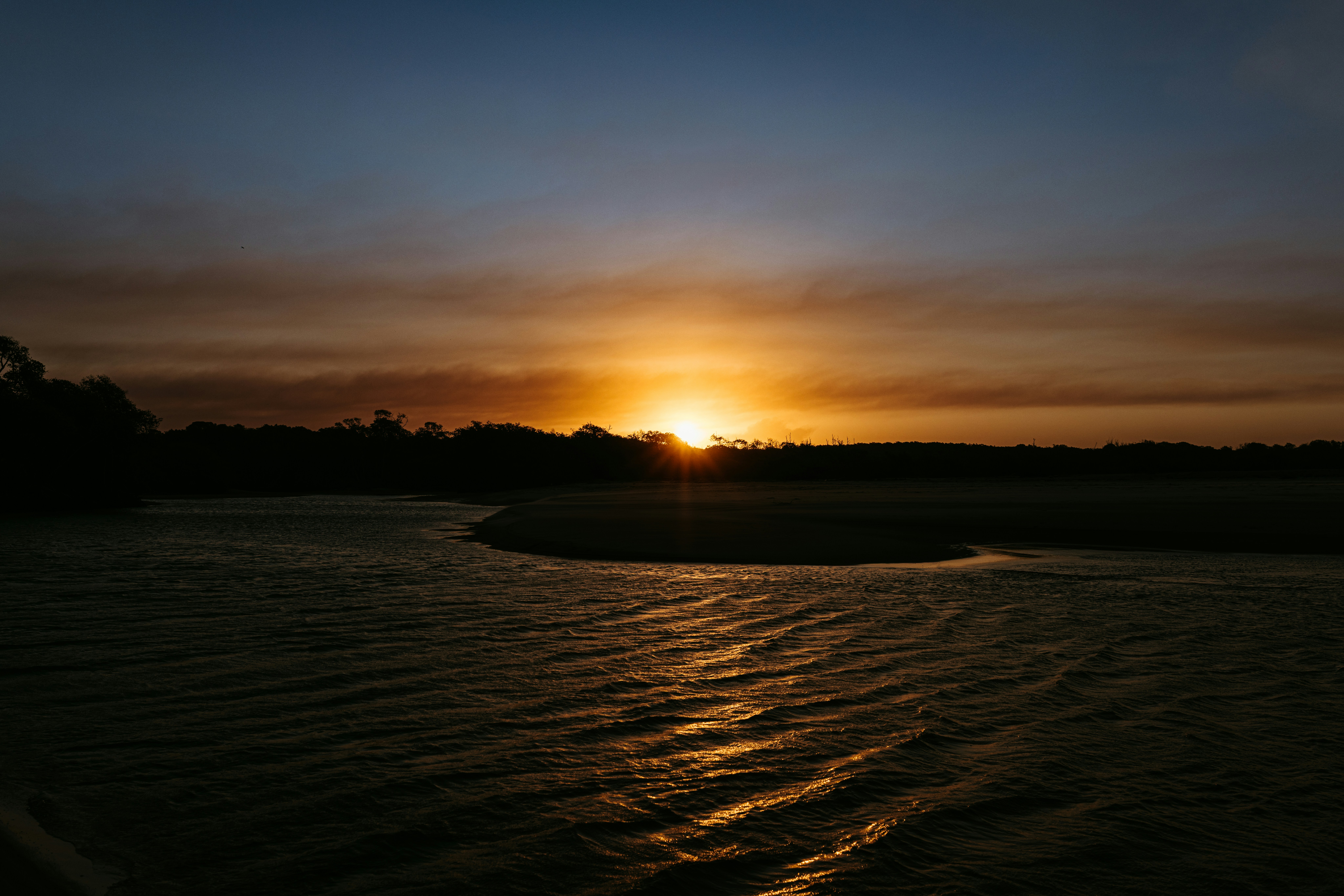 Sol bajo sobre un río oscuro con una línea de árboles en silueta y agua ondulante
