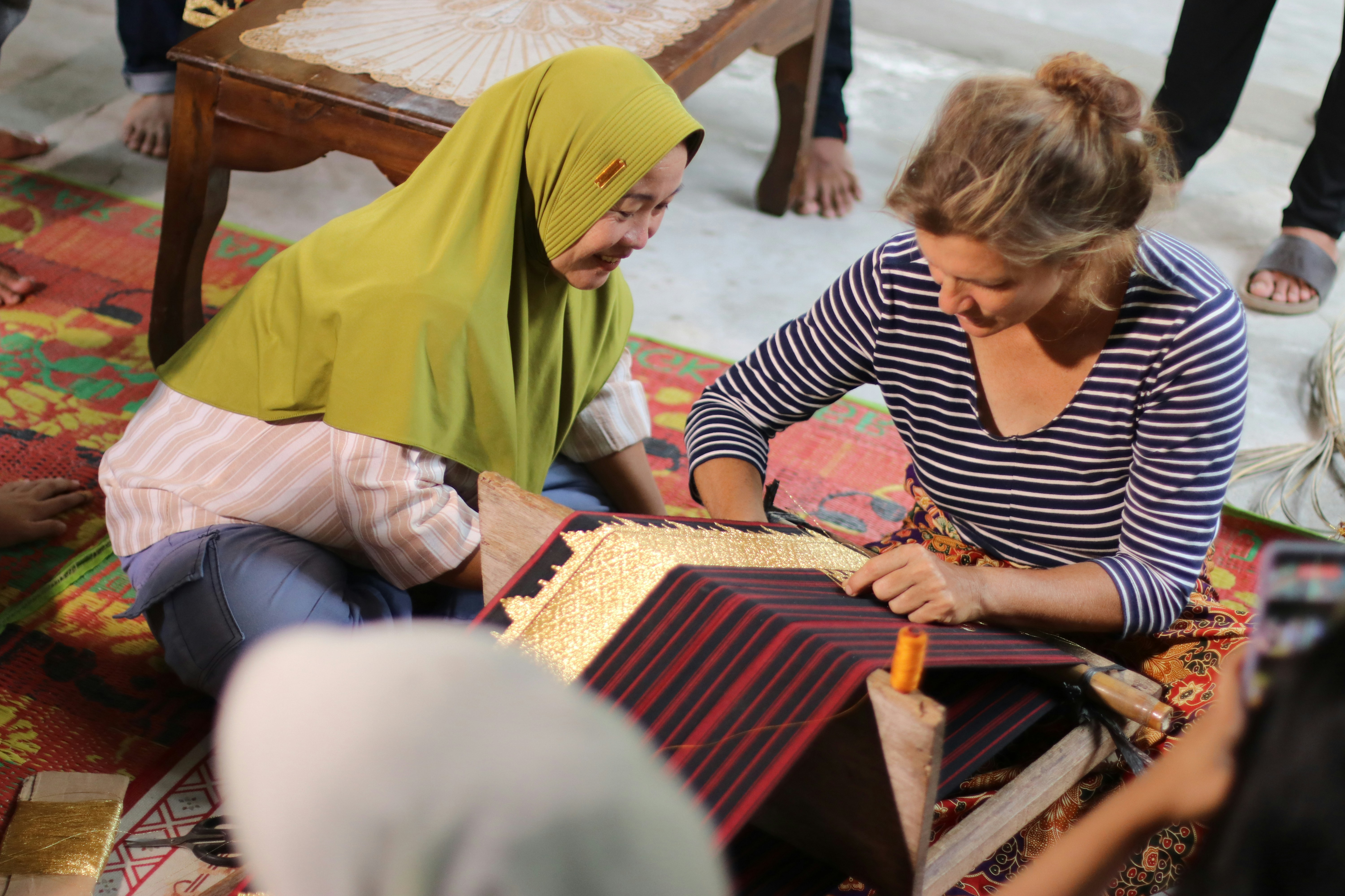Zwei Frauen, die ein gemustertes Textil auf einem kleinen Holzwebstuhl auf einer Matte weben