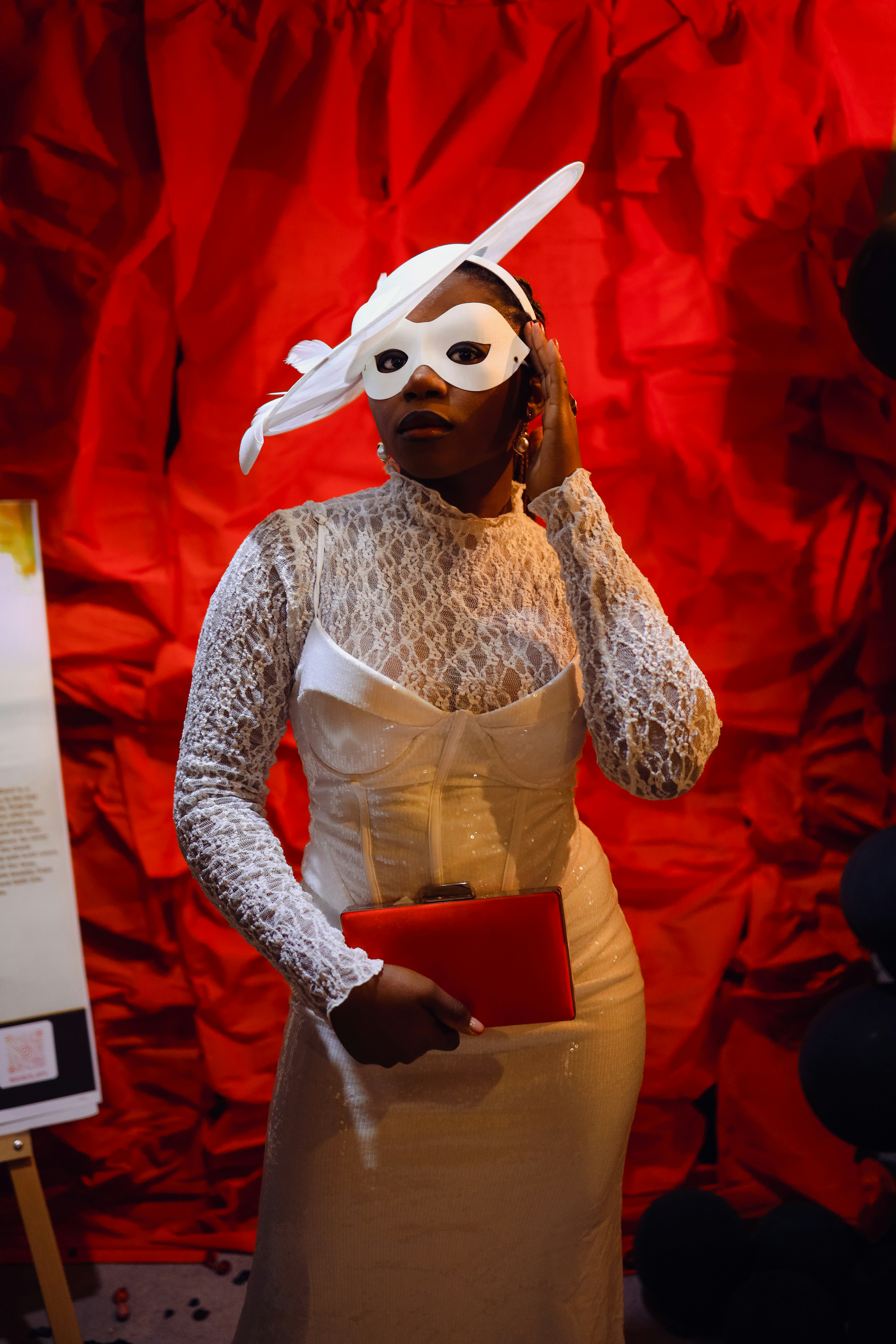 A person in a white lace dress and mask holding a red clutch