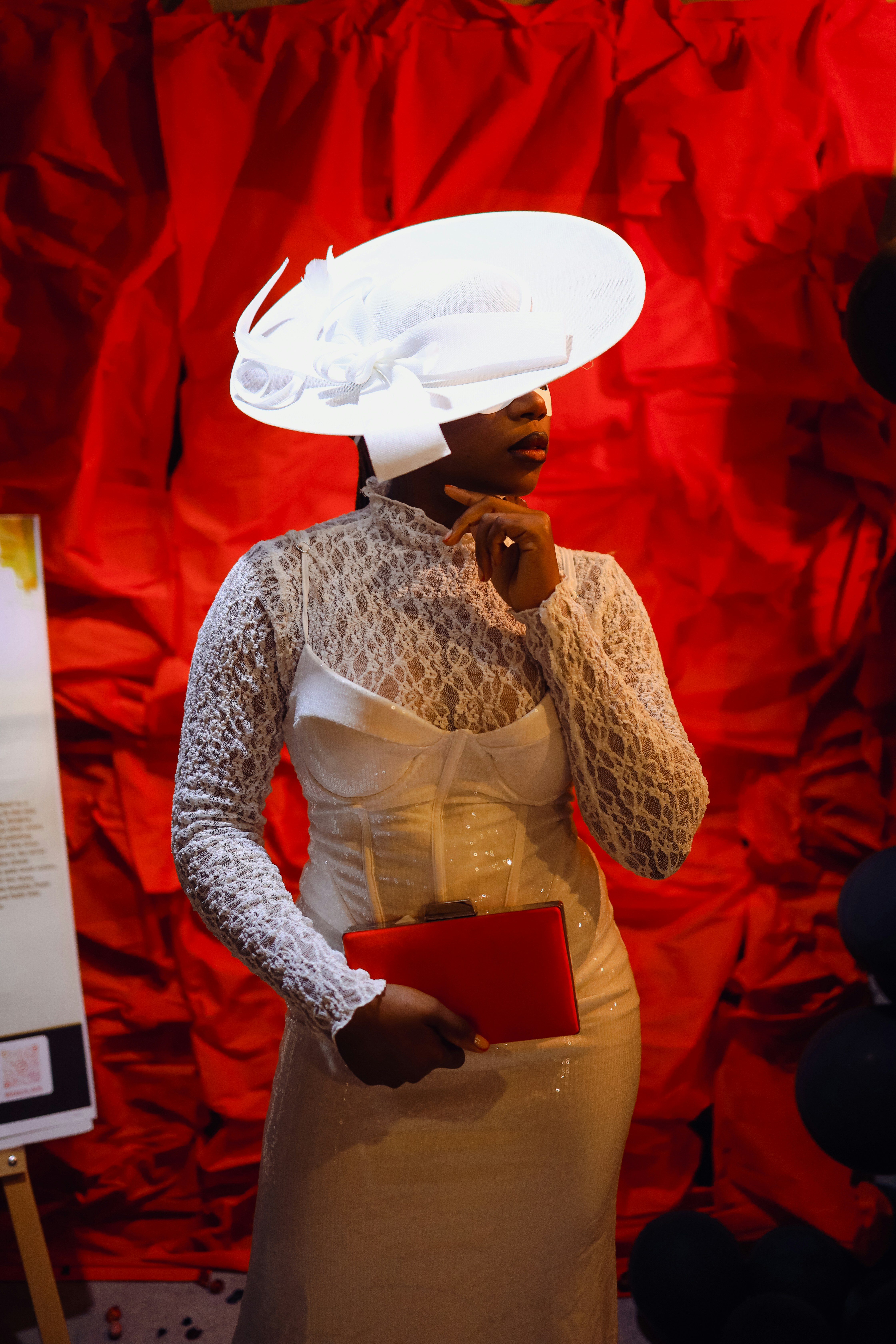 A woman in a white lace dress and wide-brimmed hat holding a red clutch
