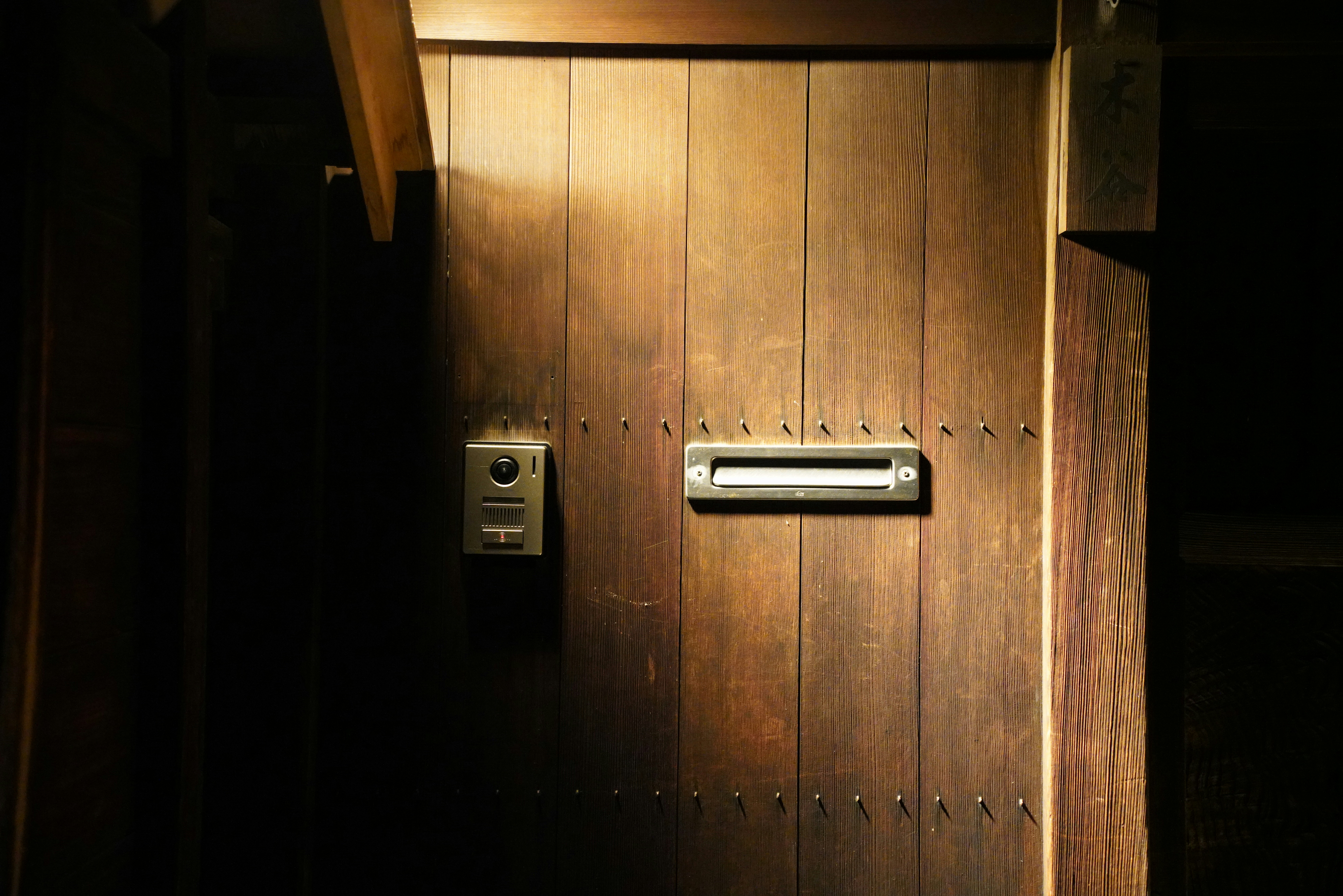 Painel de interfone com câmera numa porta de madeira, ao lado da fenda de correio, sob uma faixa de luz.