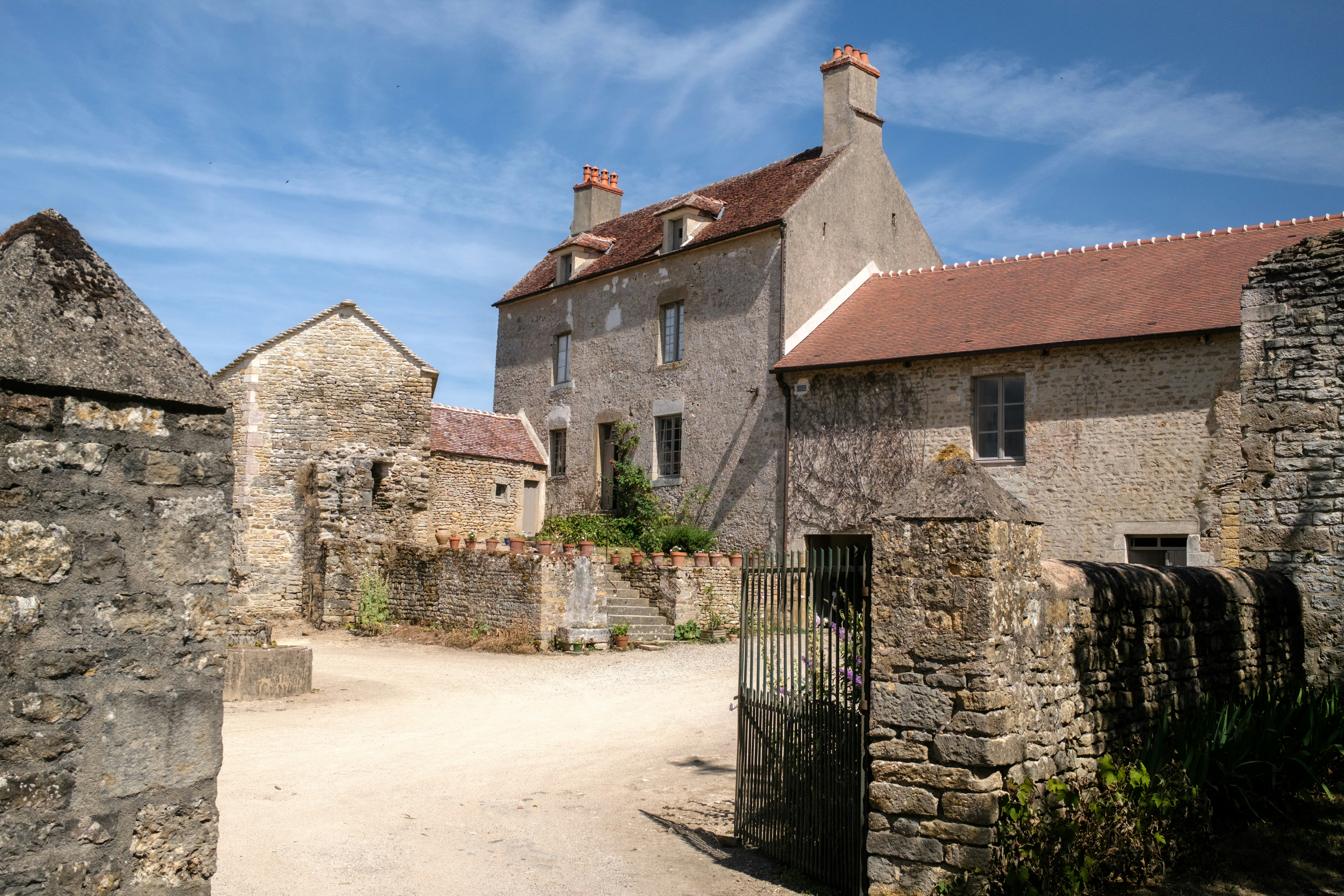 Corps de ferme en pierre aux toits de tuiles, avec un portail en fer ouvert sur une cour de terre battue sous un ciel bleu