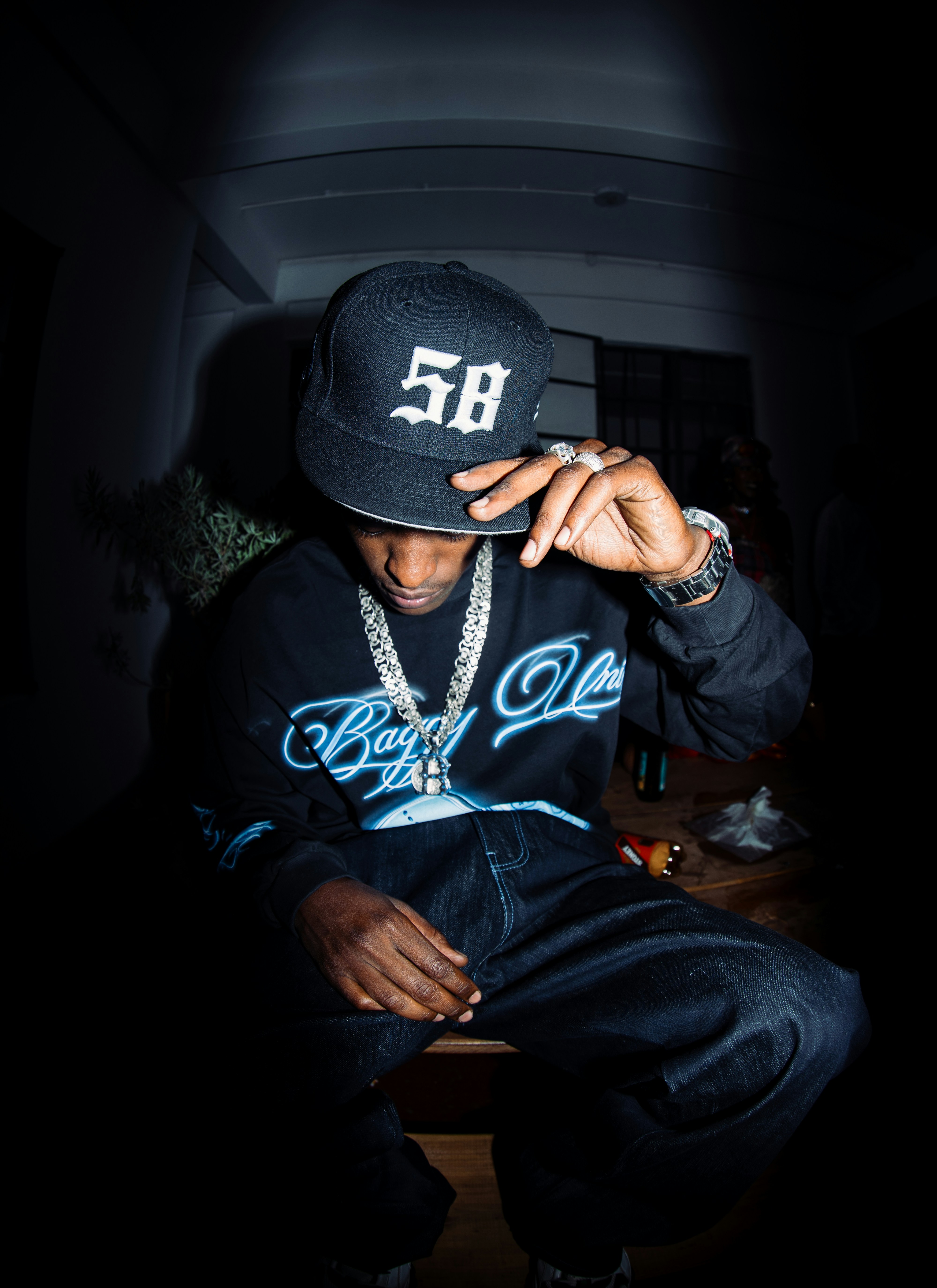 A person in a black cap and sweatshirt with silver chains in dim light