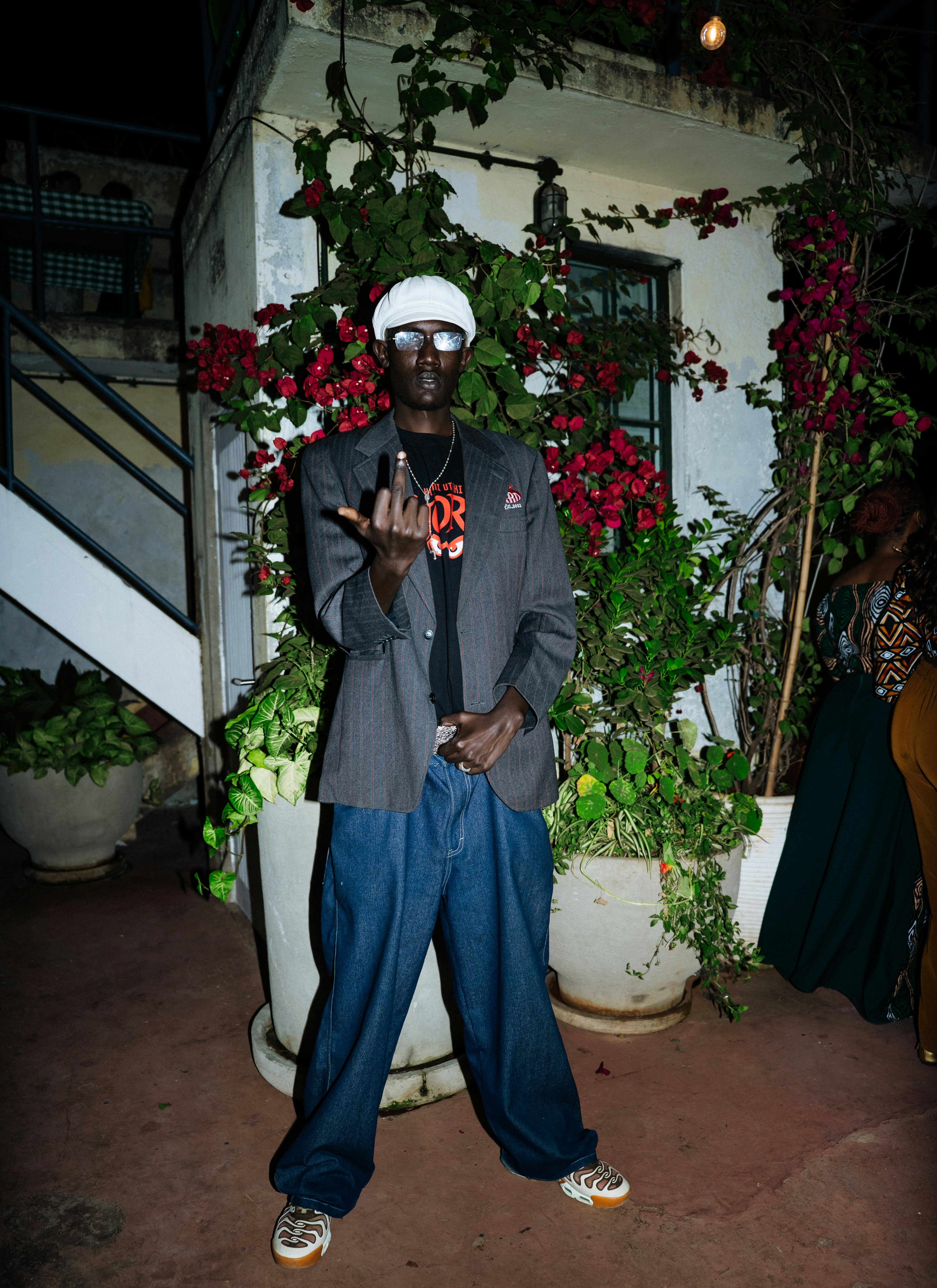 A person in a white cap and grey blazer gesturing with a middle finger
