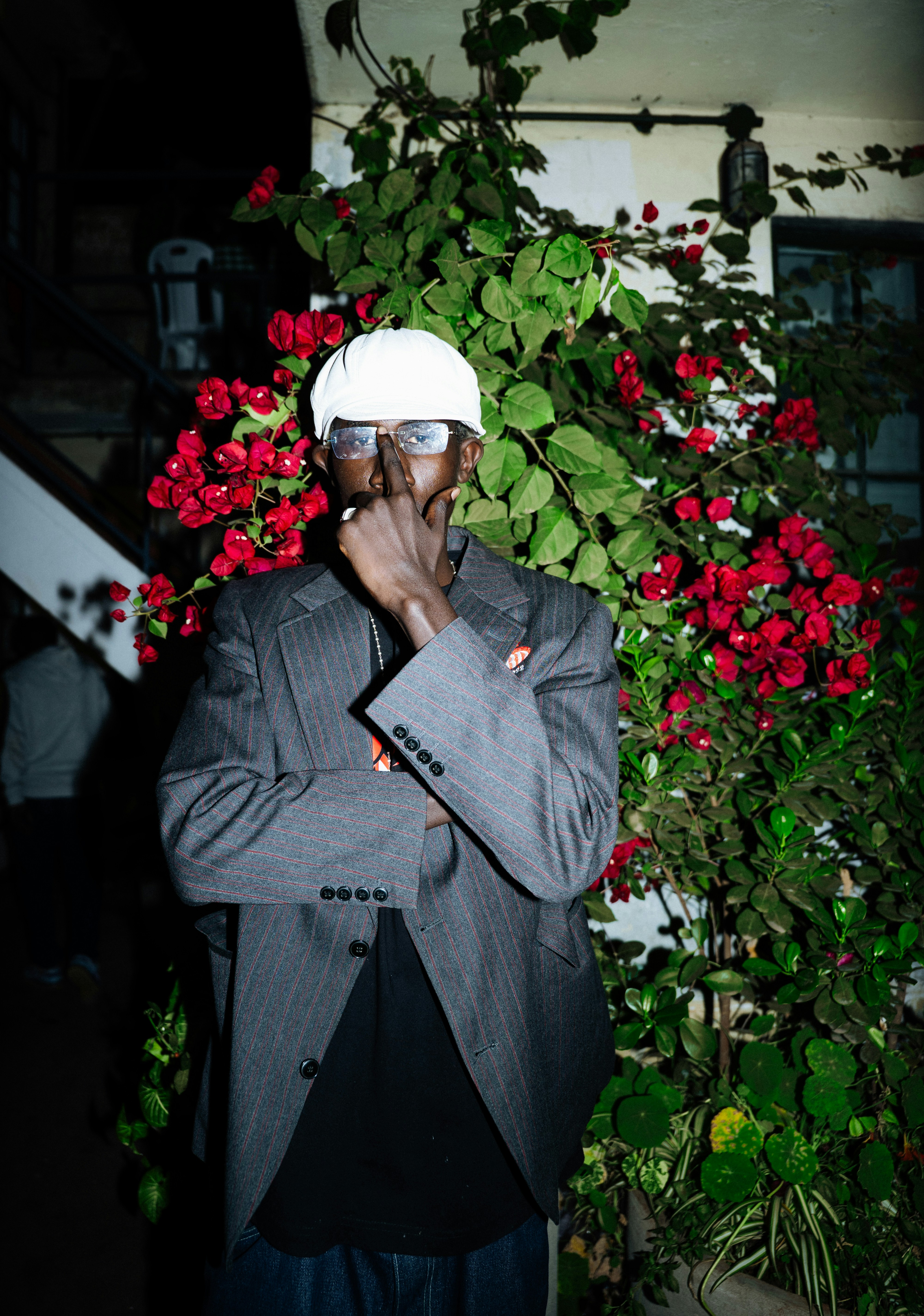A person in a white cap and pinstripe blazer standing before red bougainvillea flowers