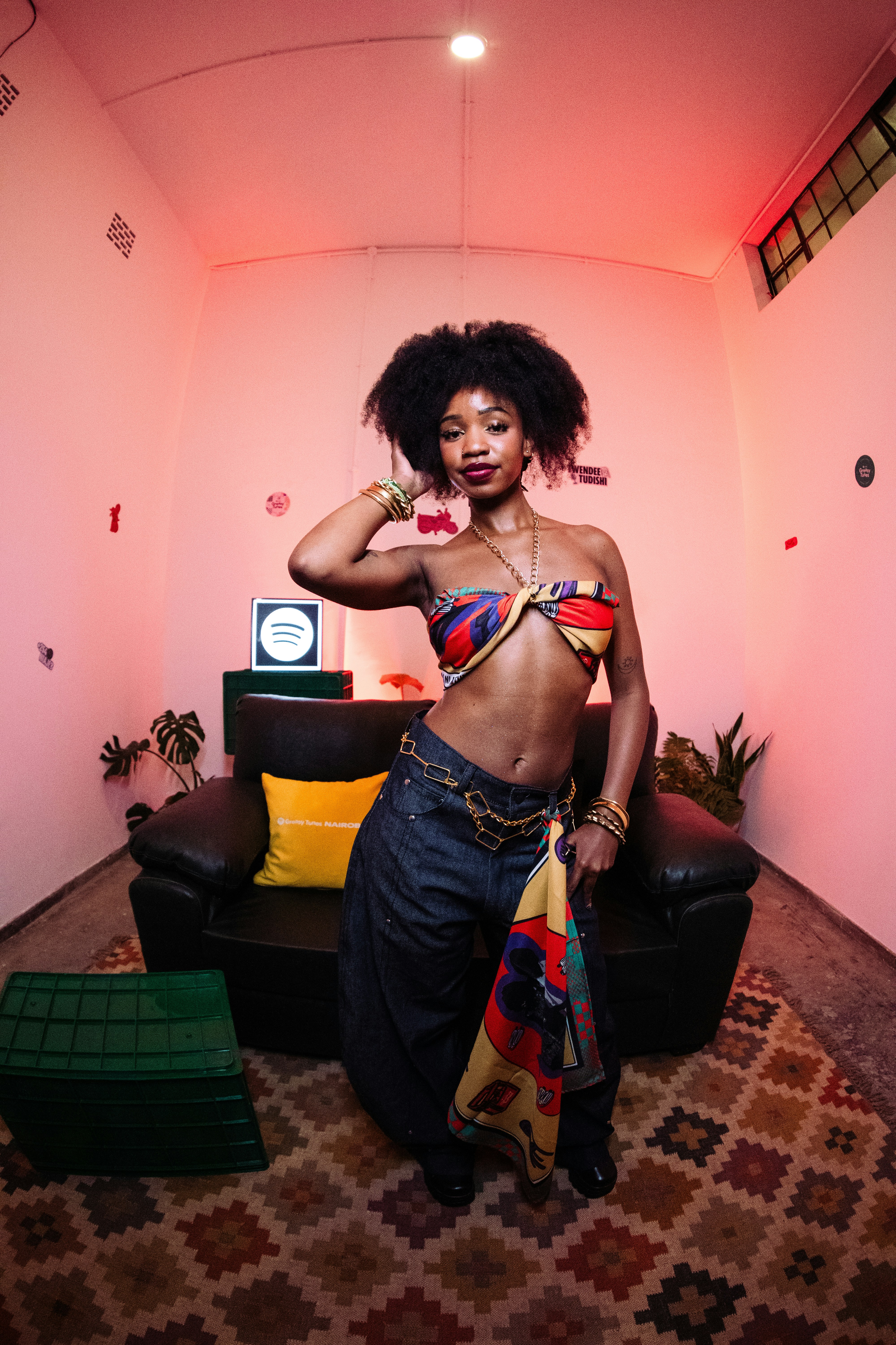 A woman with an afro wearing a patterned scarf top and jeans in a room