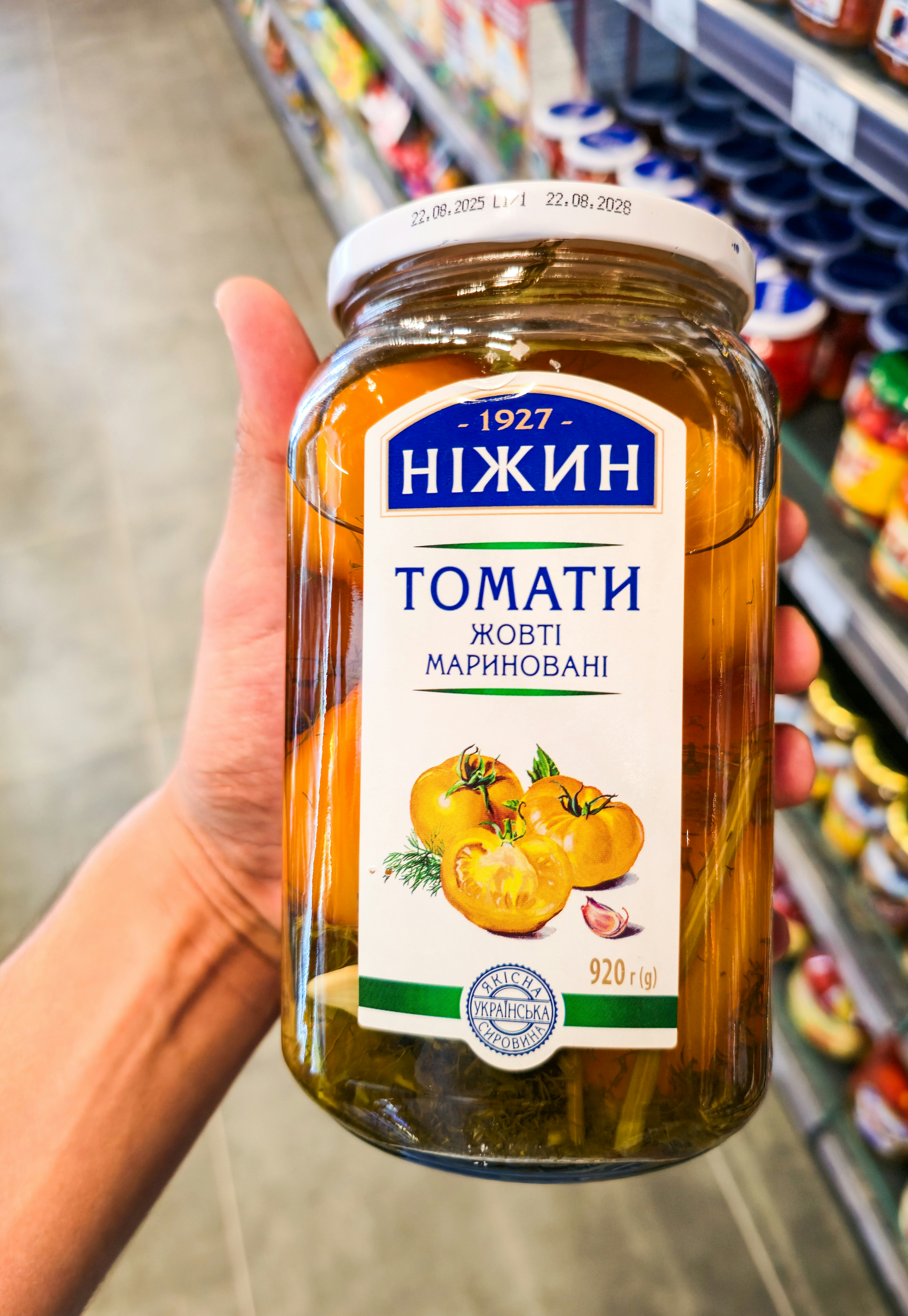 A hand holding a glass jar of pickled yellow tomatoes in a grocery store