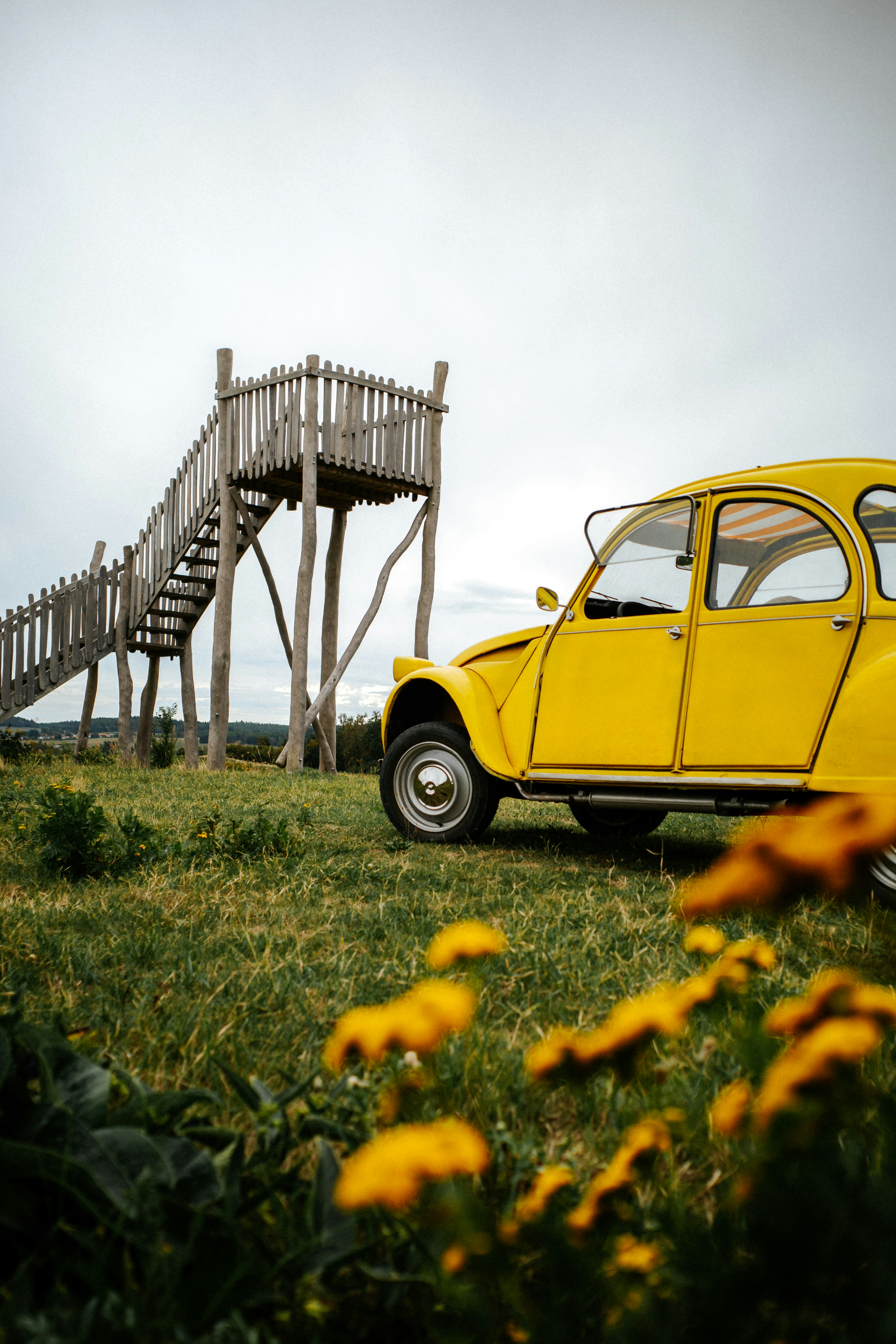 Una Citroën 2CV gialla parcheggiata su un campo erboso vicino a una torre di osservazione in legno