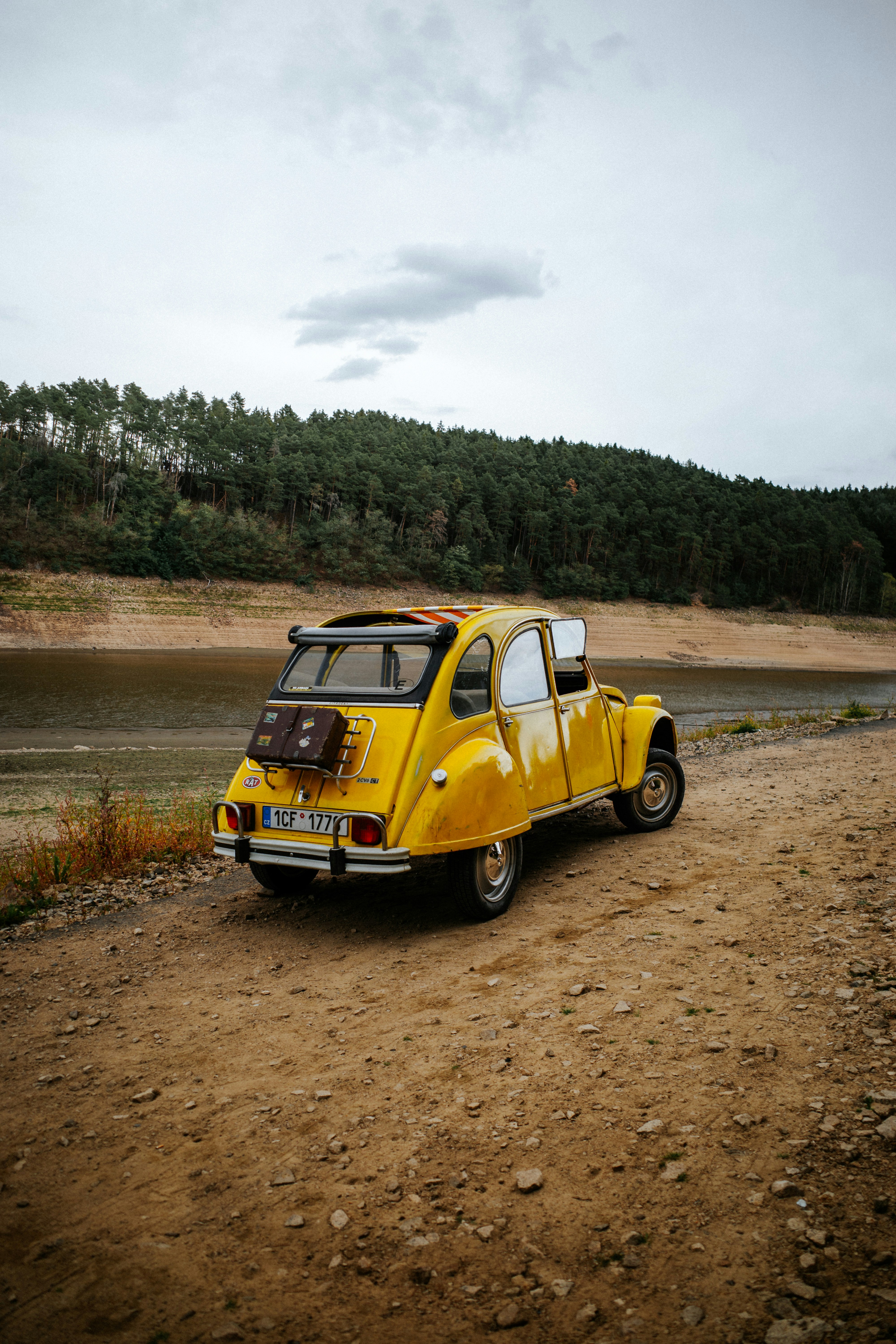Una Citroën 2CV gialla parcheggiata su un sentiero sterrato accanto a un fiume e a una foresta