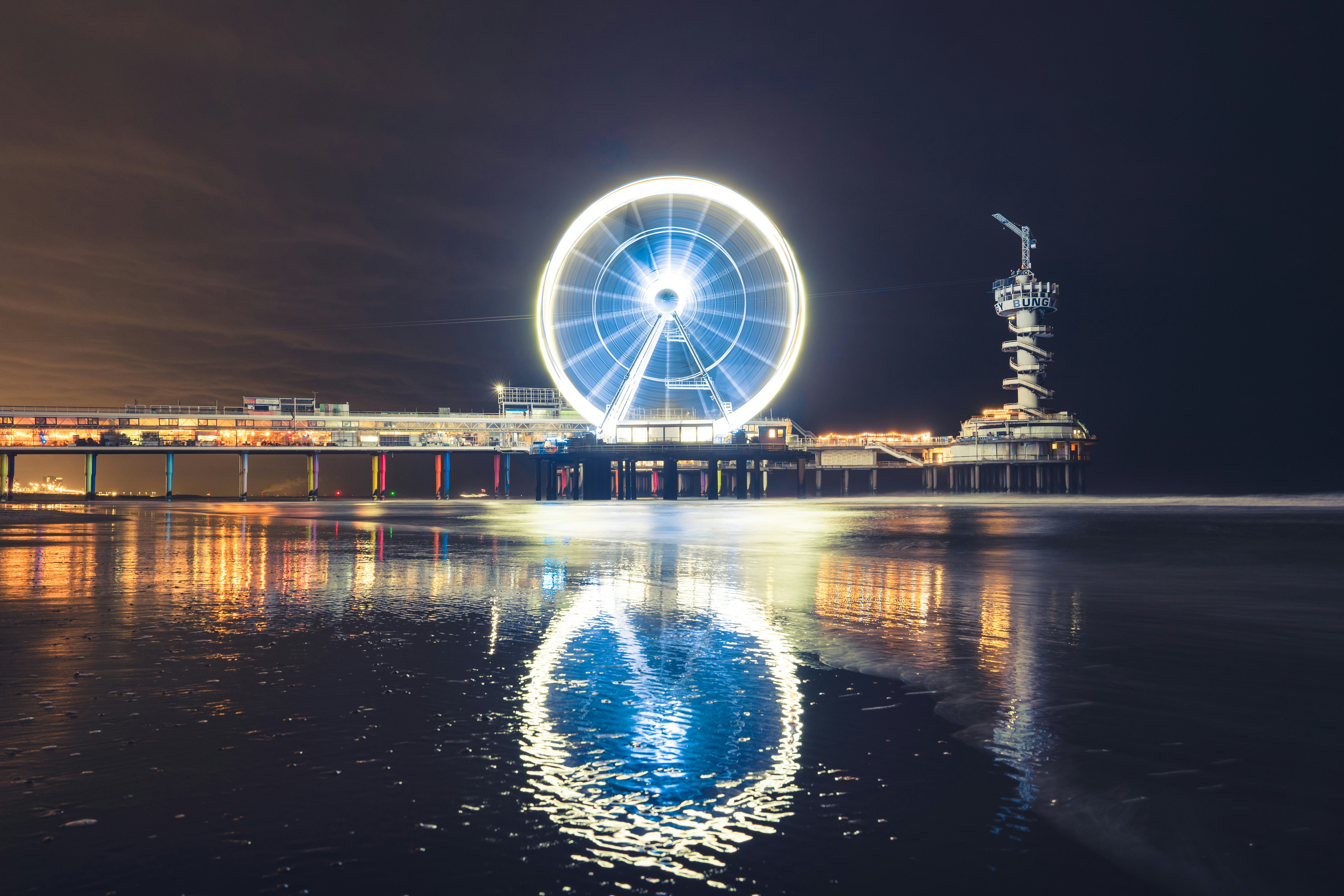 Uma roda-gigante brilhante em um píer refletindo na areia molhada à noite