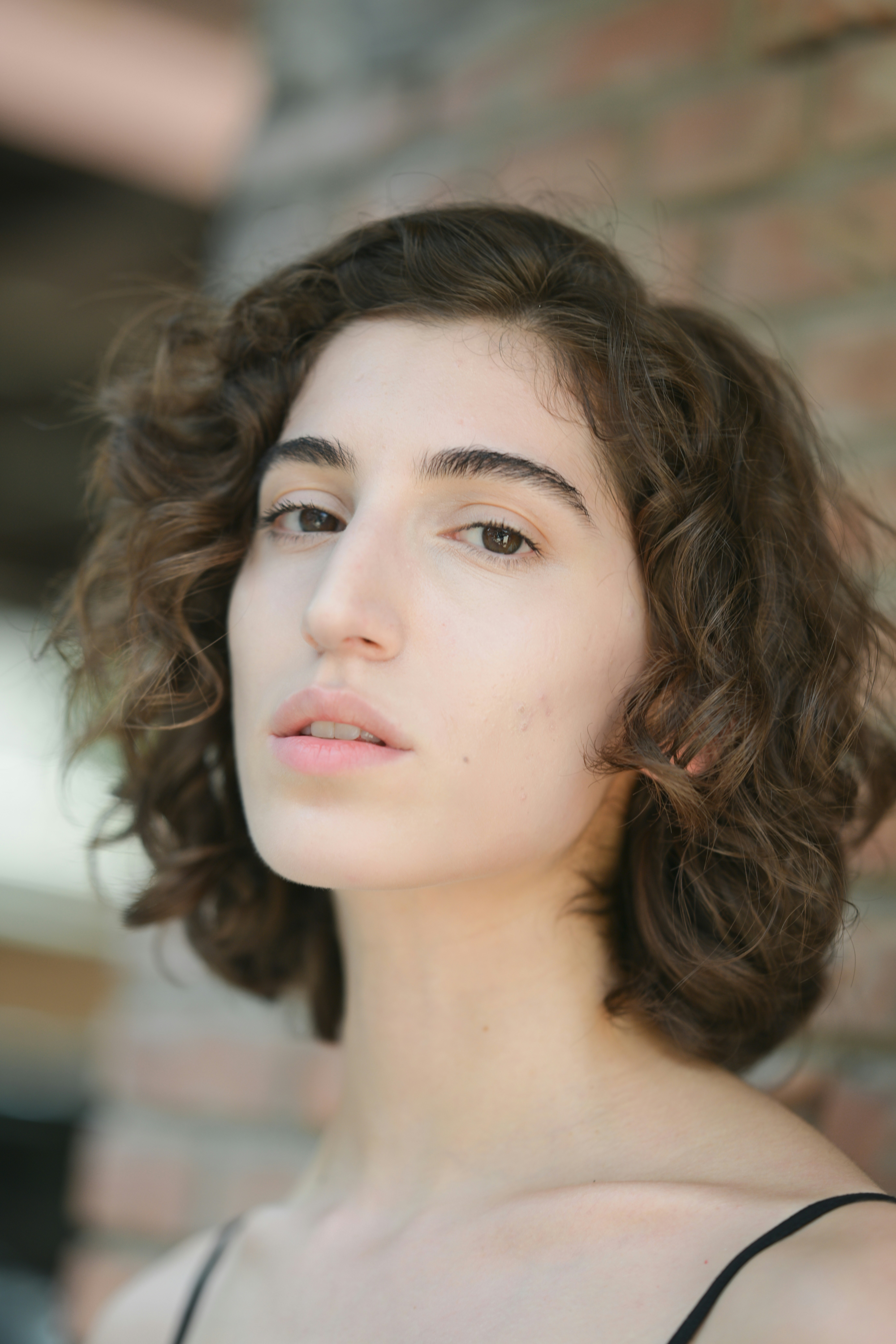 A woman with short curly brown hair against a blurred brick wall