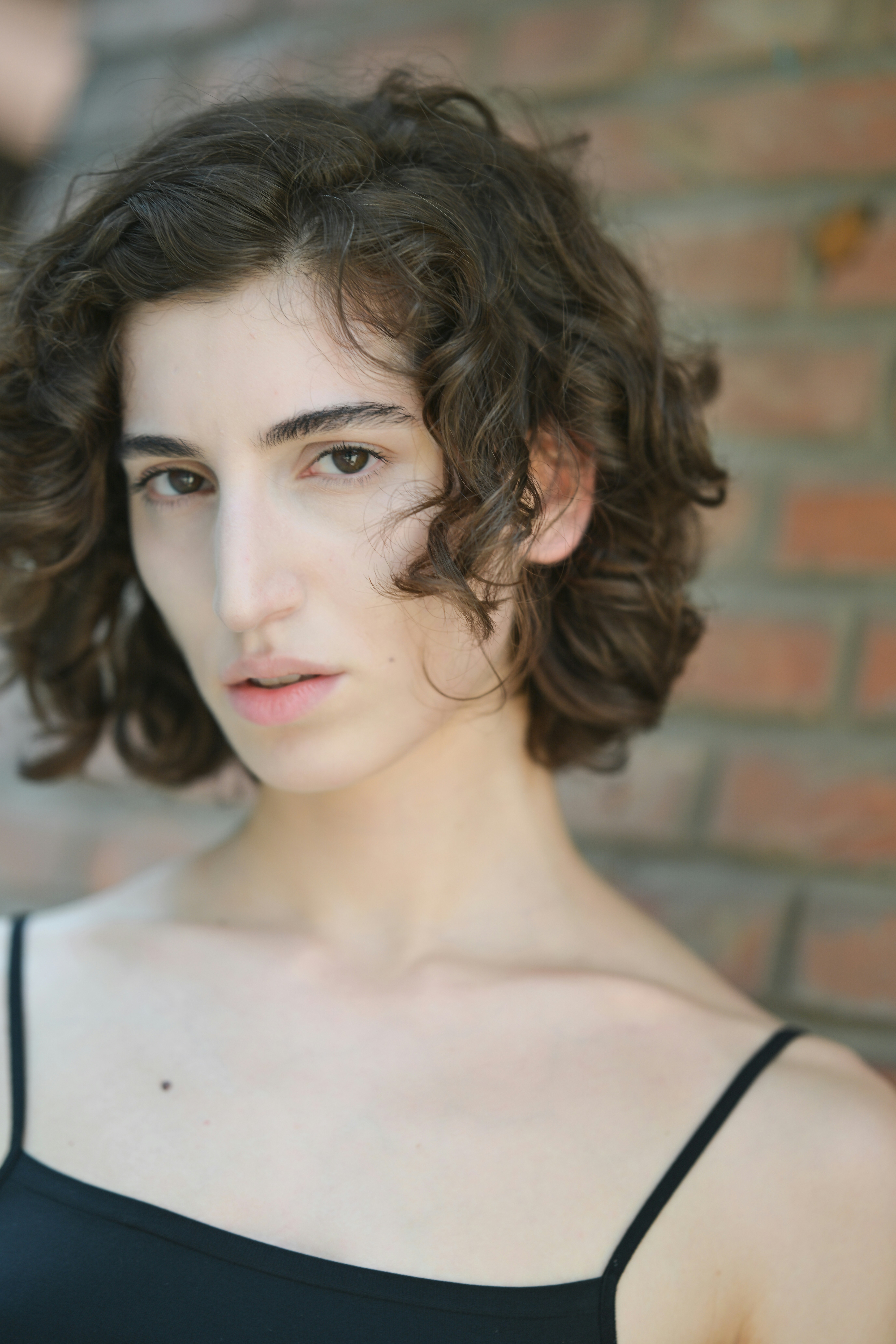 A woman with curly brown hair in a black top against a brick wall