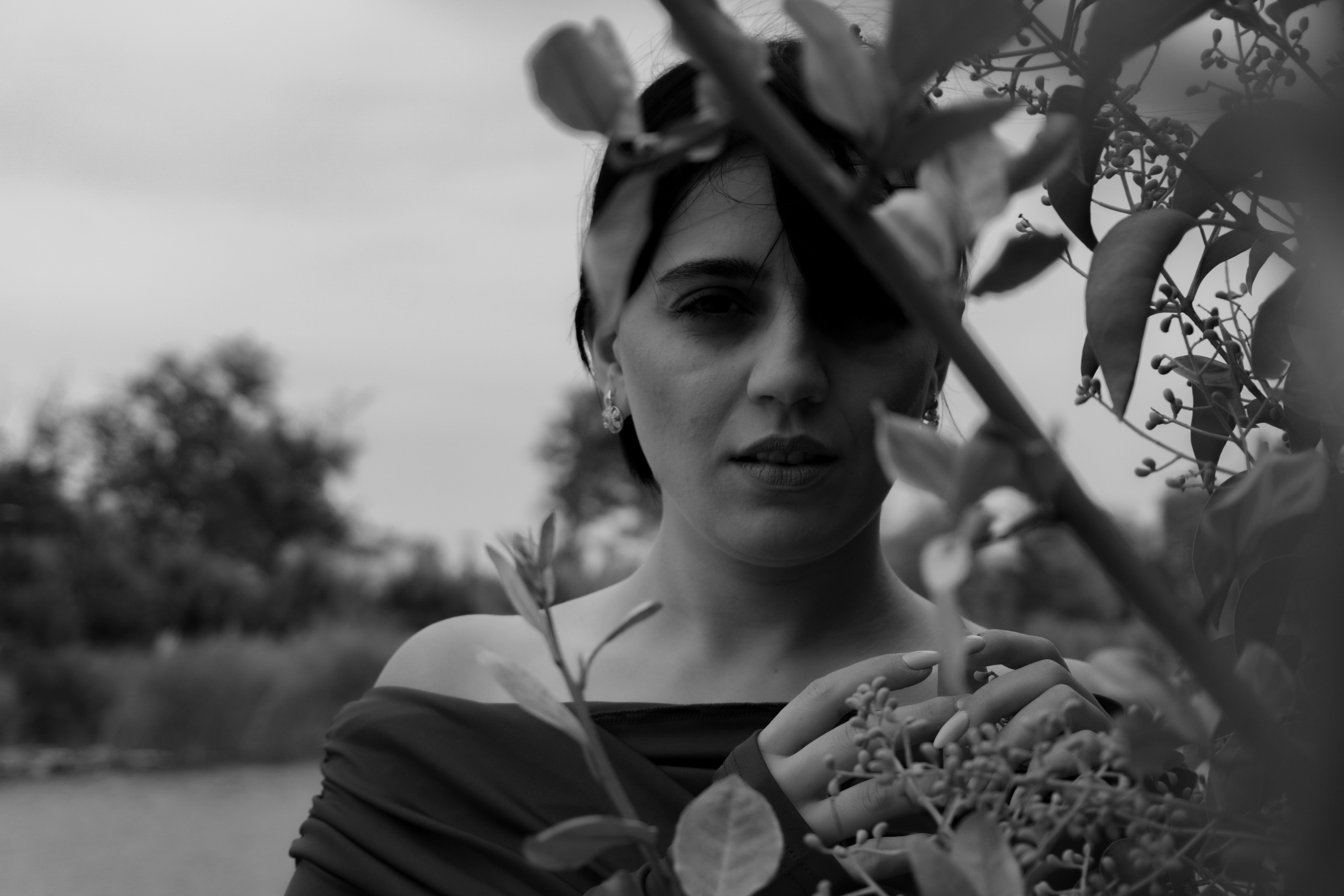 A woman in an off-the-shoulder top behind leafy branches in black and white