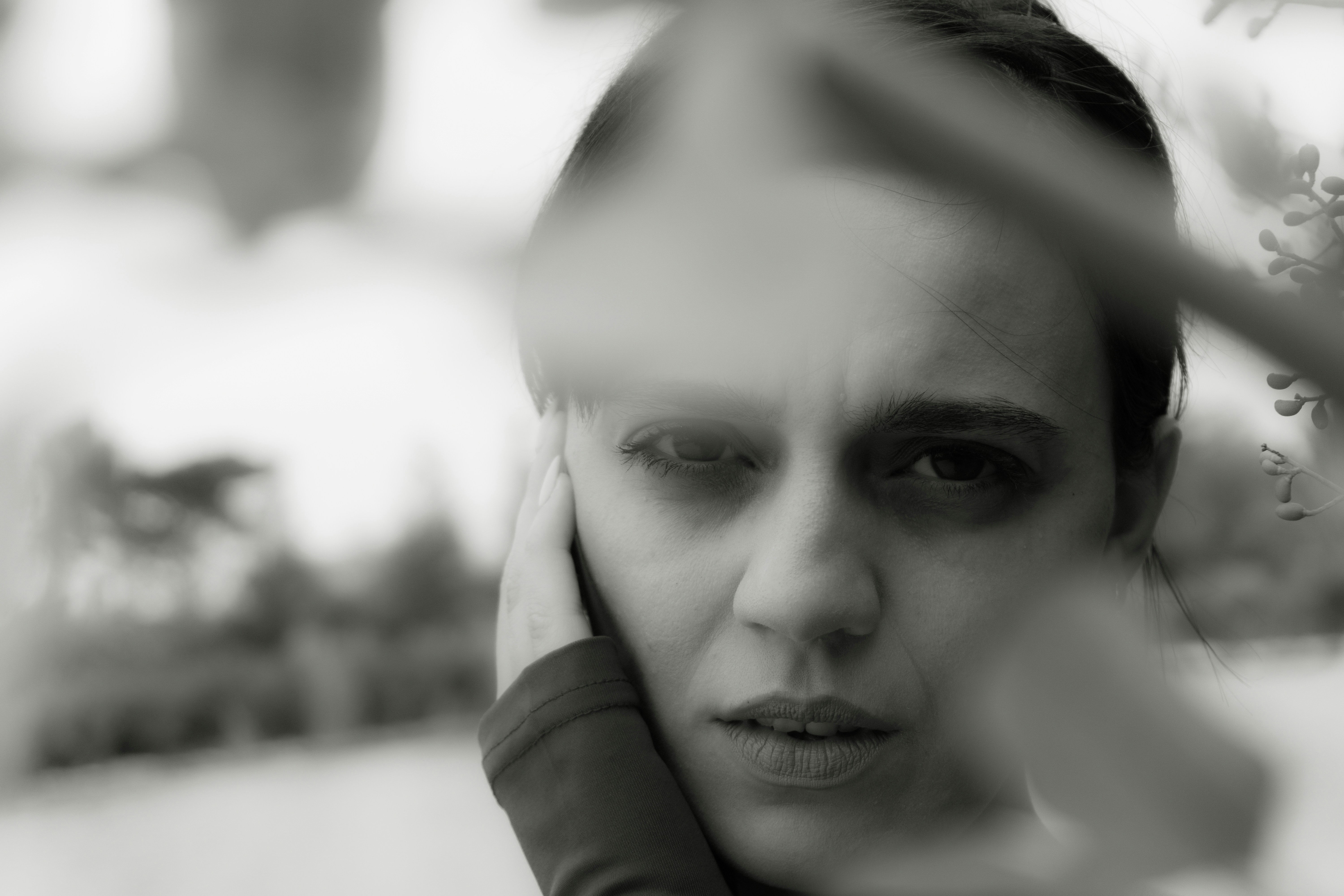 A woman with her hand against her cheek, viewed through blurred foreground foliage