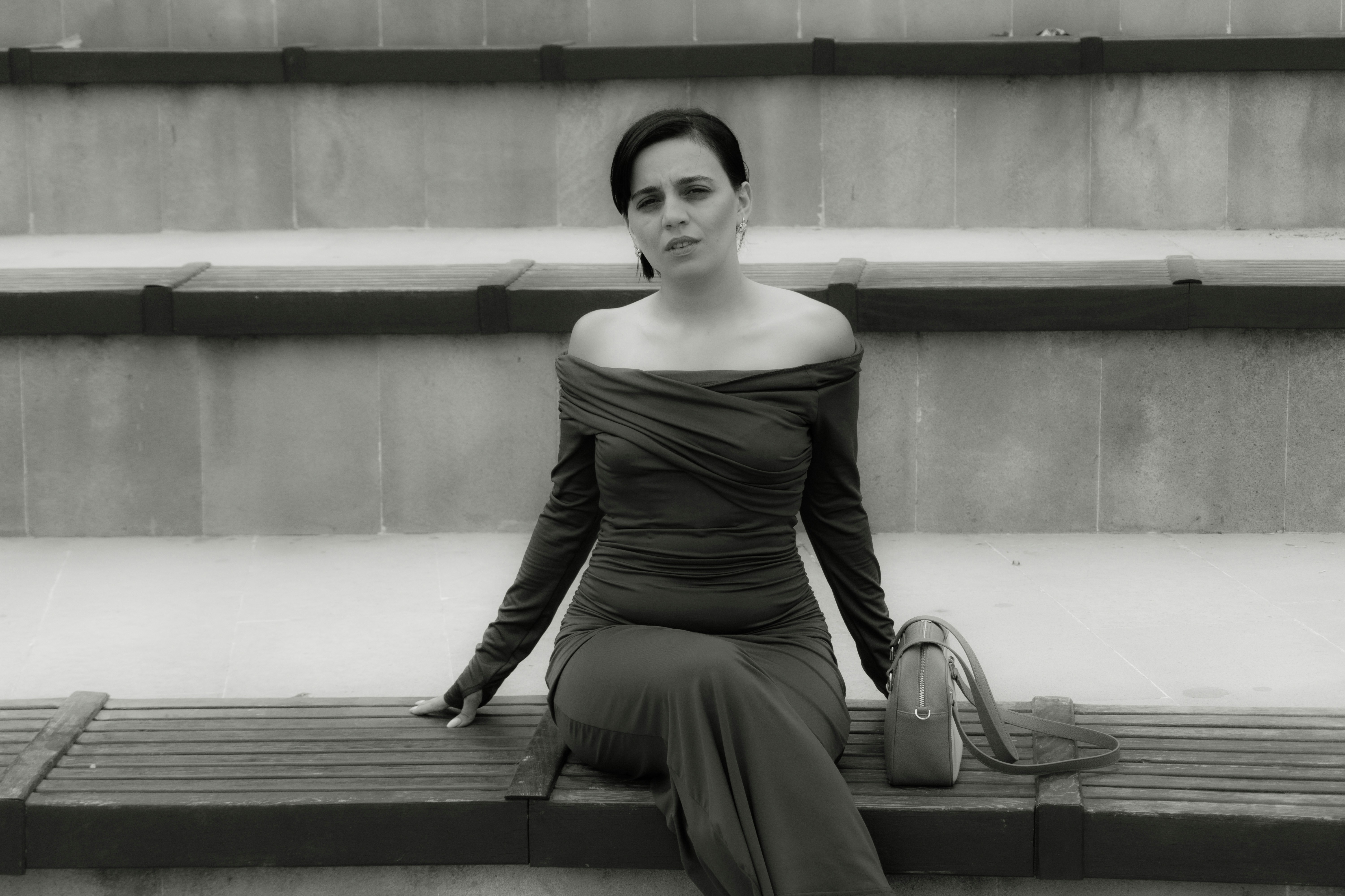 A woman in an off-the-shoulder dress on a wooden bench with a handbag