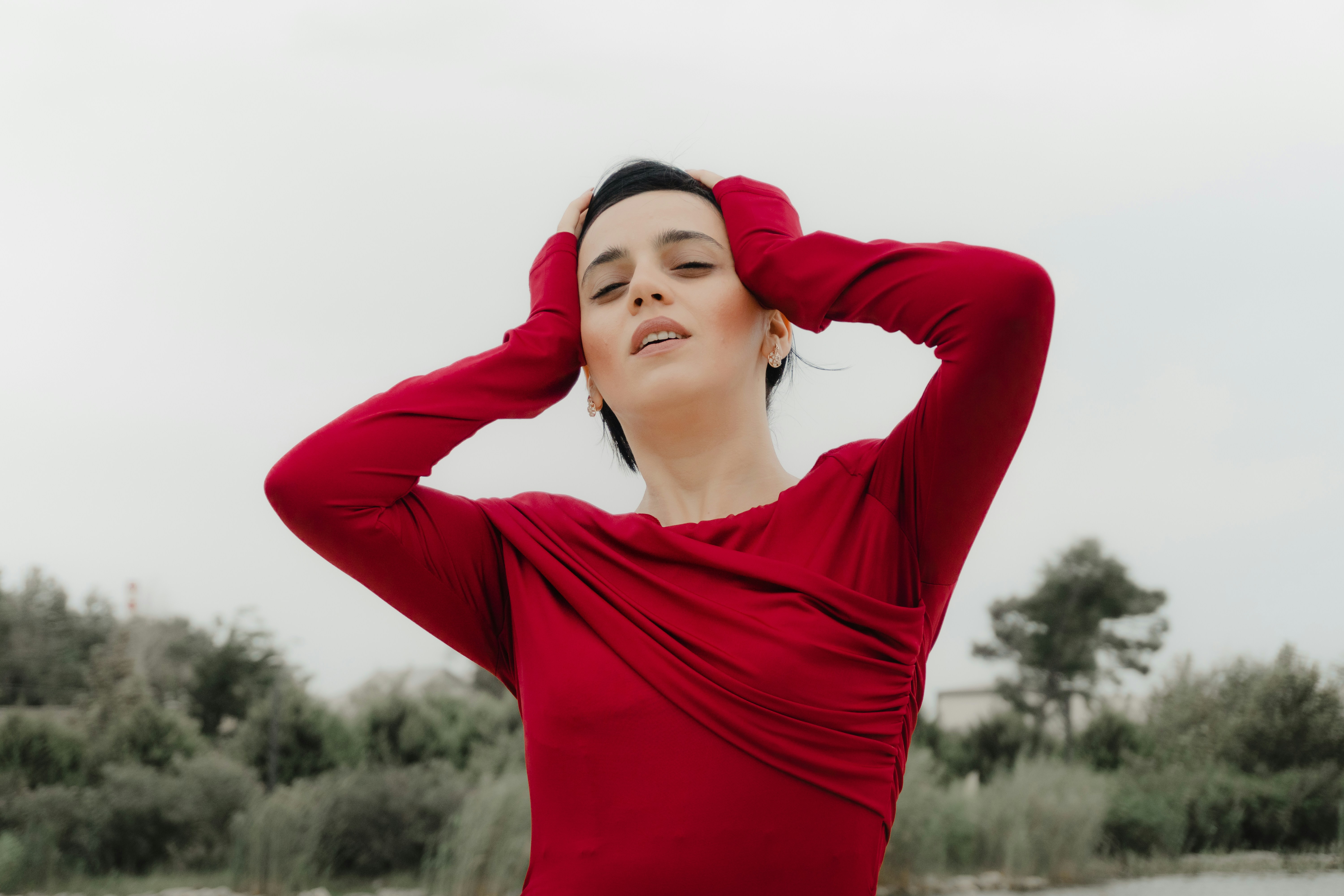 A woman in a red long-sleeved dress with hands on her head outdoors