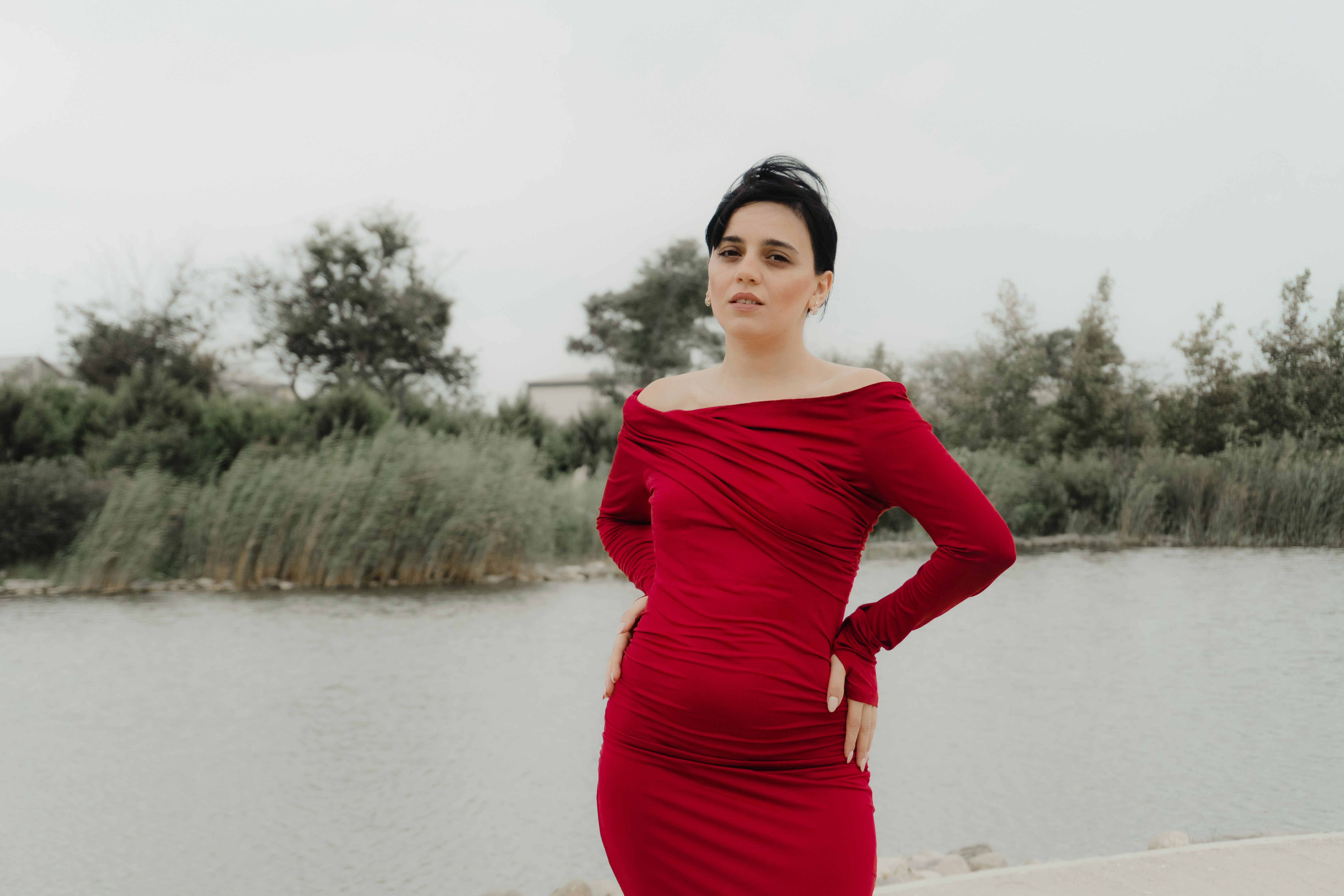 A woman in a long-sleeved red dress standing by a lake with reeds